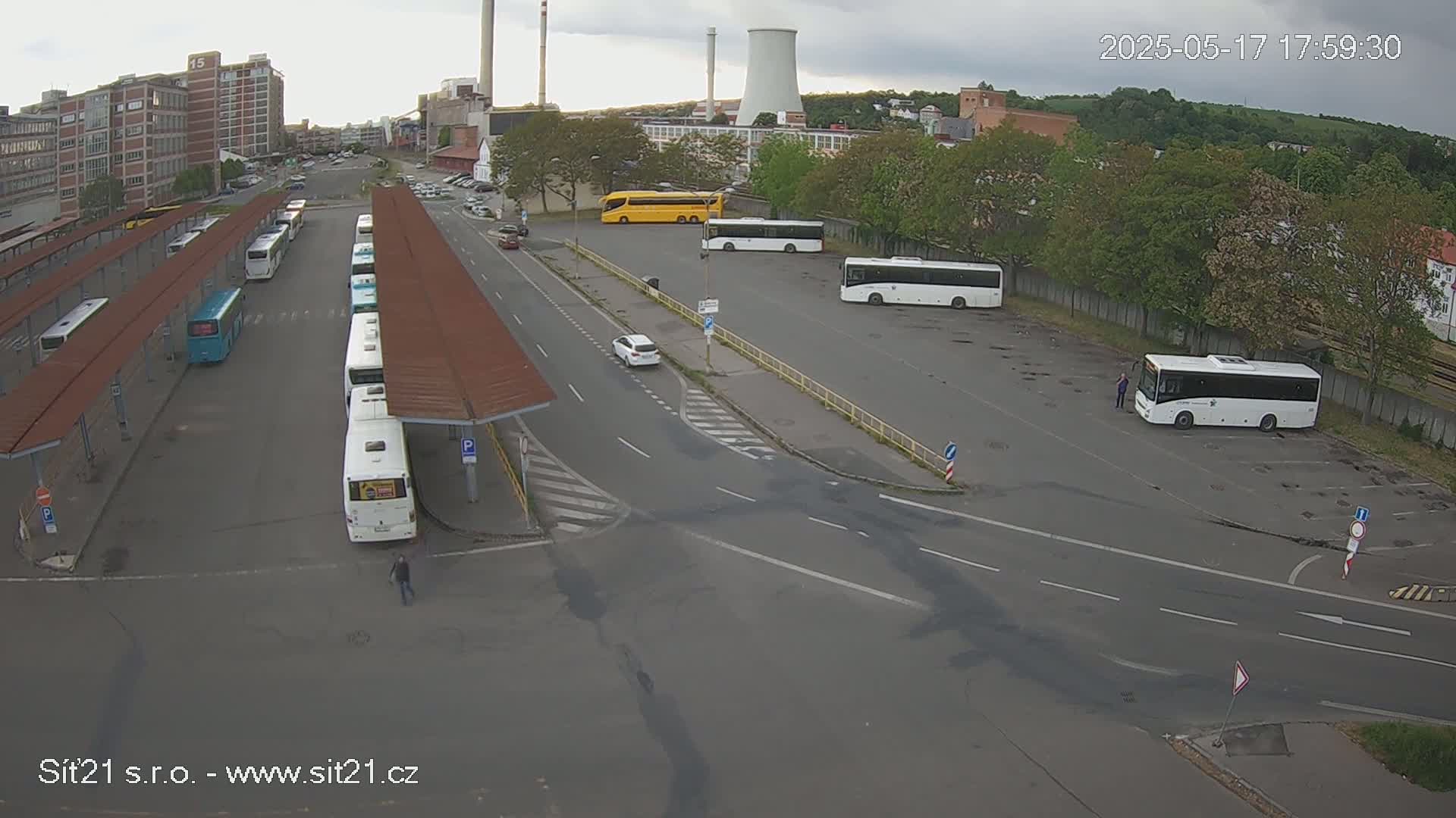An overcast day reveals a bus station with numerous buses parked under shelters and in a nearby lot, along with a few cars and a person walking.