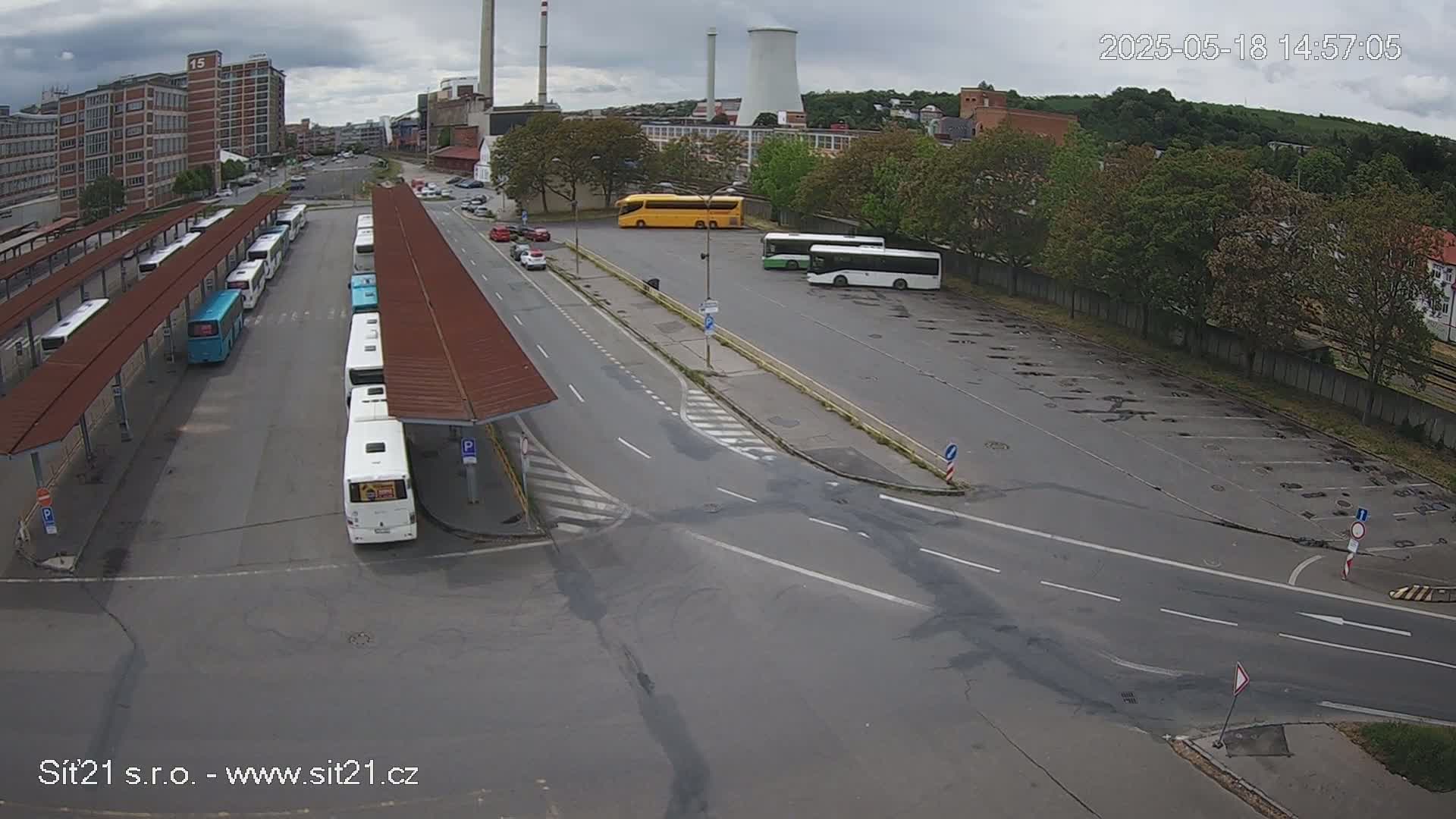 A bus station with many buses parked under covered shelters, some additional buses parked in a nearby lot, and an overcast sky.