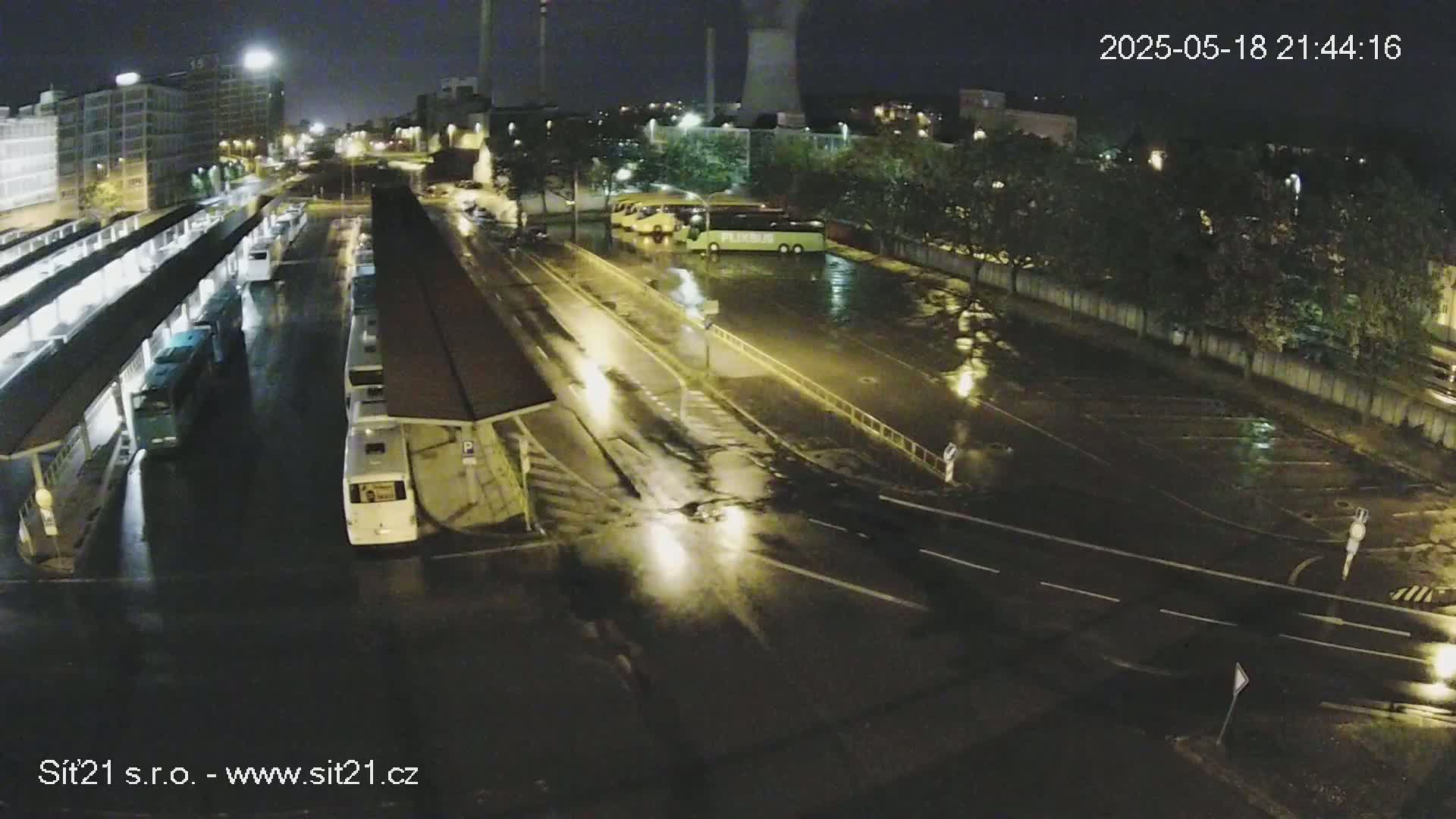 A nighttime, rain-soaked bus station with several buses parked under shelters and a few cars on the surrounding wet roads.