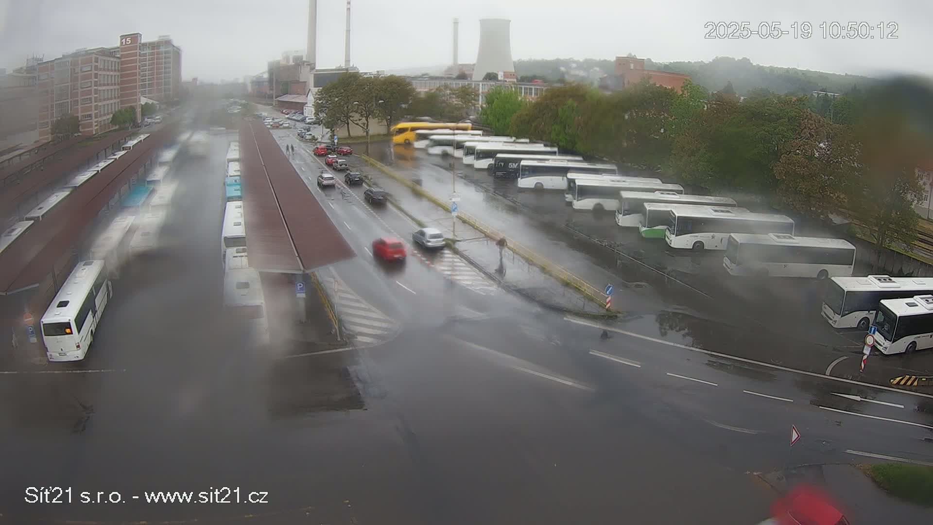 A rainy day view of a bus station with numerous buses parked under covered areas and in a lot, with several cars traveling on adjacent roads.