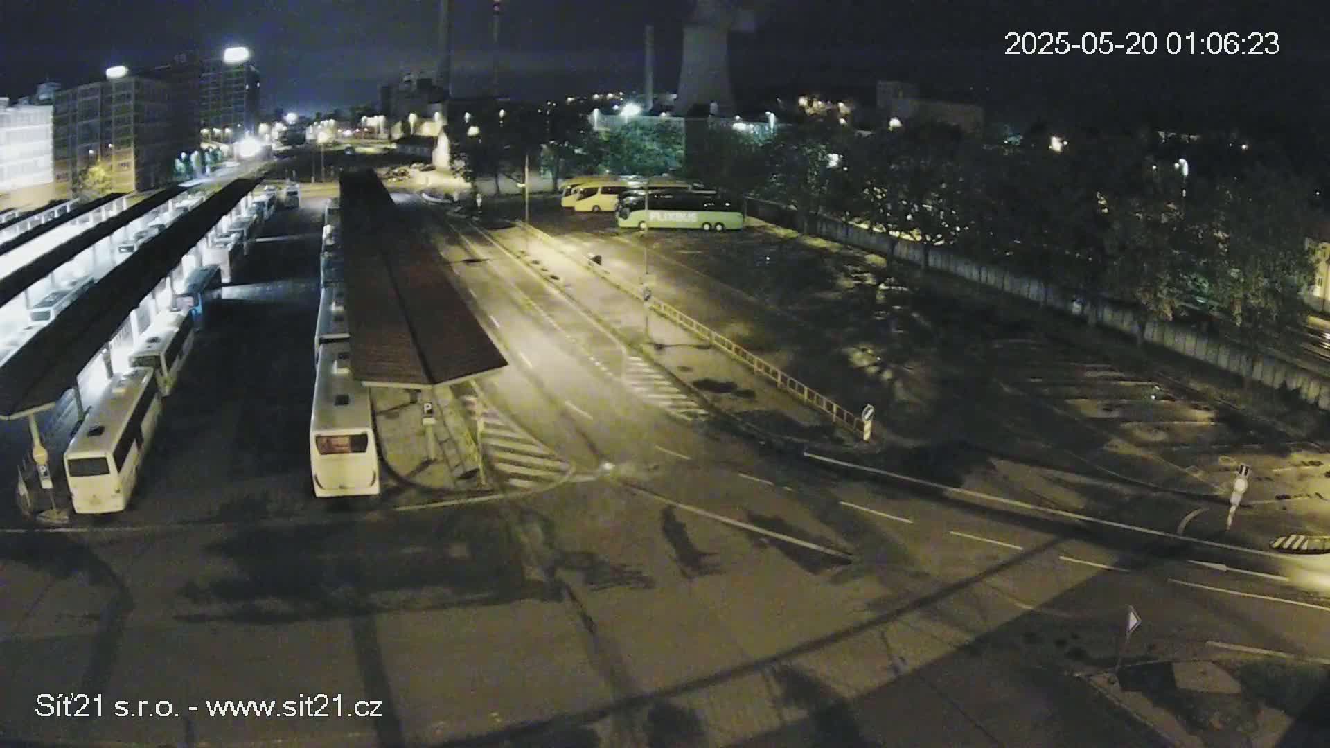 A nighttime high-angle view shows a bus station with numerous buses parked under shelters, adjacent to a road with several other buses parked nearby.