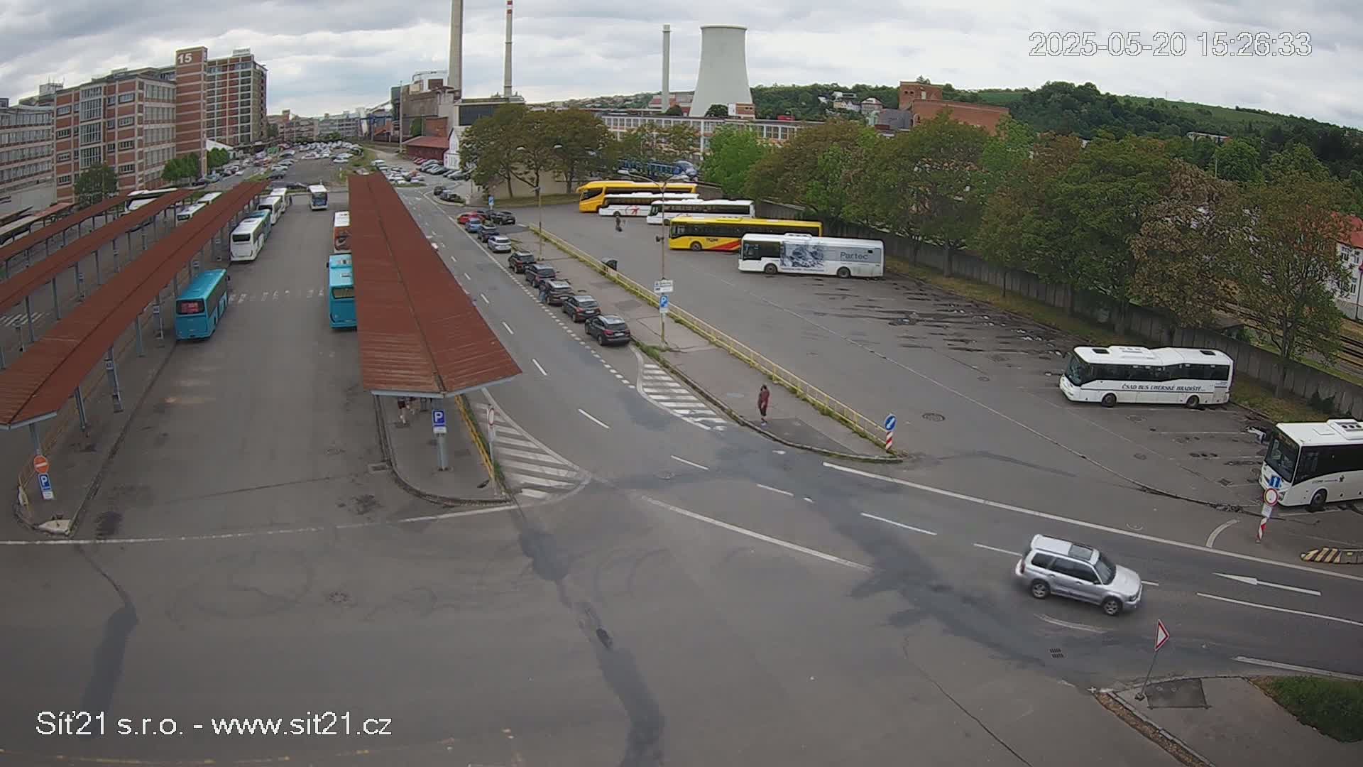 A bus station with numerous buses parked under covered areas and in an adjacent lot, a few cars in the parking lot and on the road, under an overcast sky.