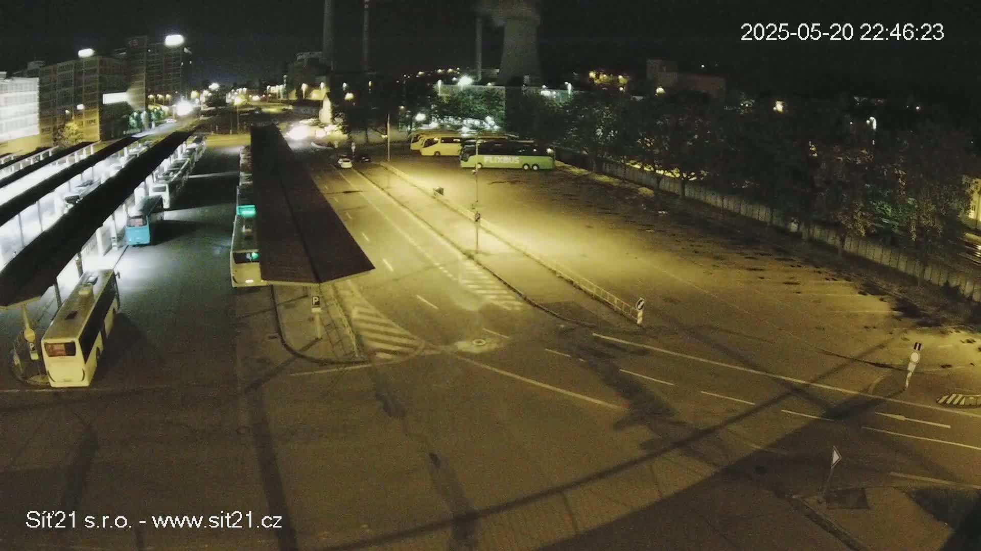 A nighttime, overhead view of a bus station with several buses parked under shelters and in a nearby lot, illuminated by artificial lights.
