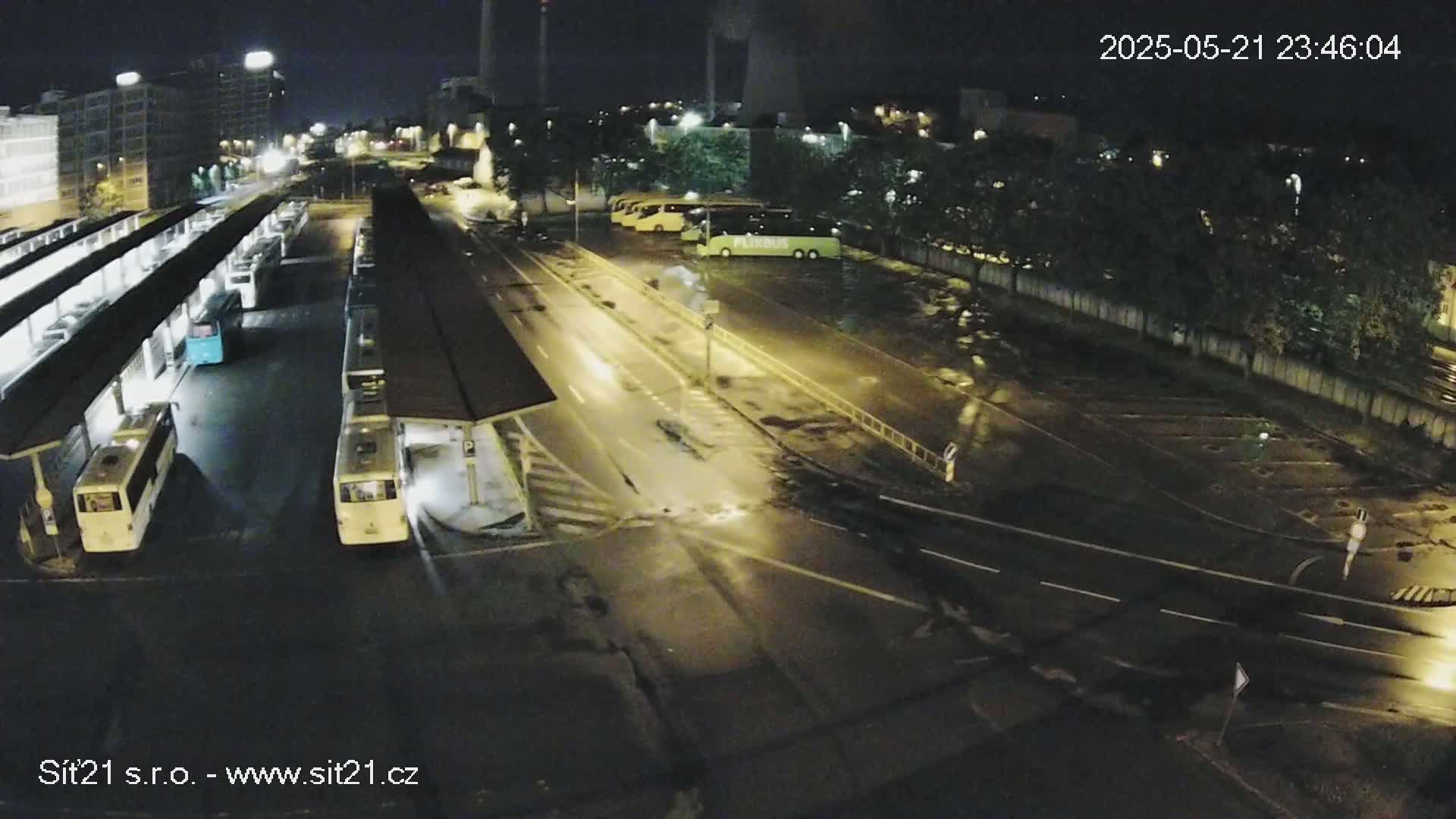 At night, under wet conditions, numerous buses are parked at a bus station, with several more parked in a nearby lot.
