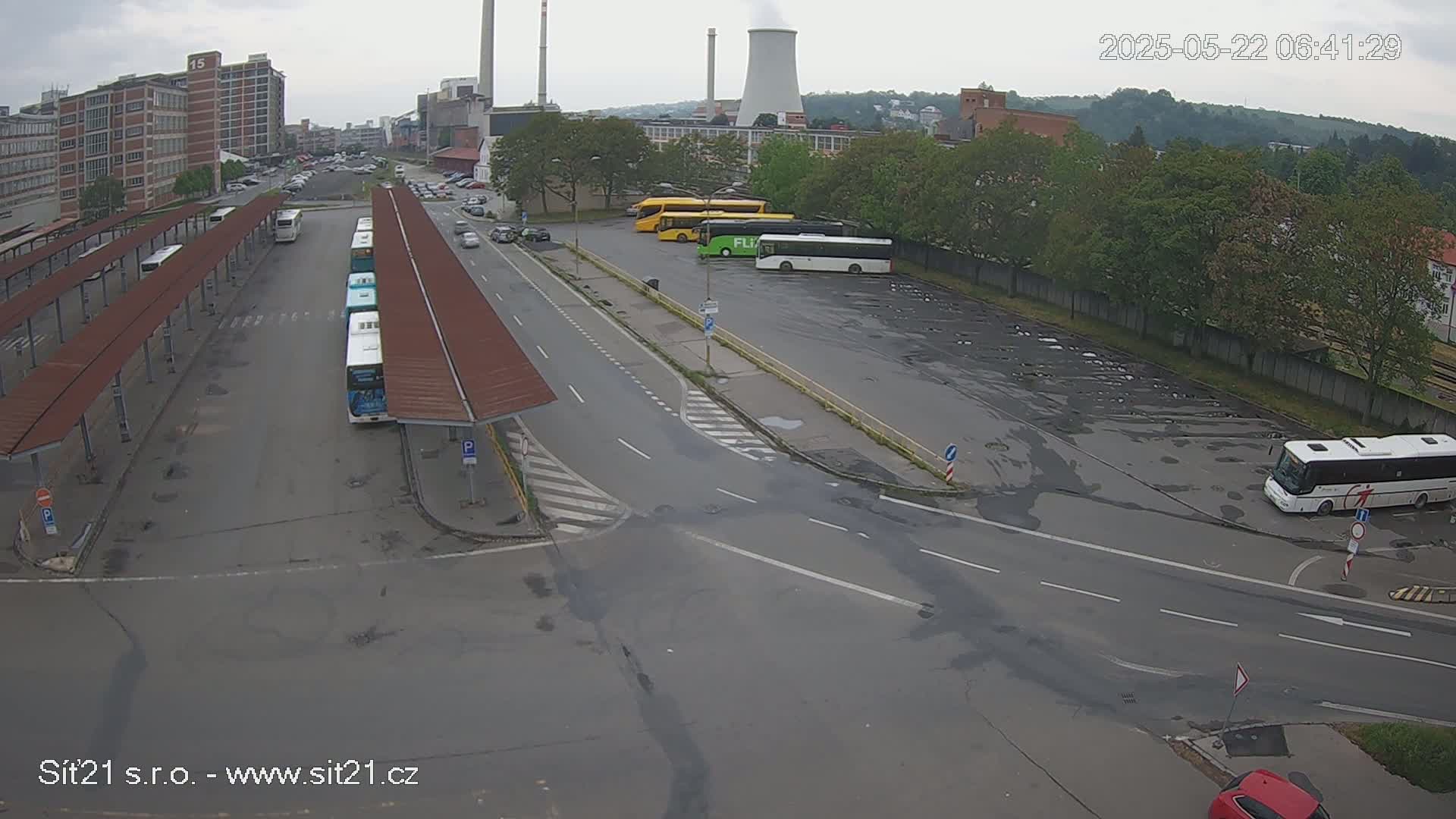 An overcast day reveals a bus station with several buses parked at shelters and in a nearby lot, next to a road intersection, and industrial buildings are visible in the background.