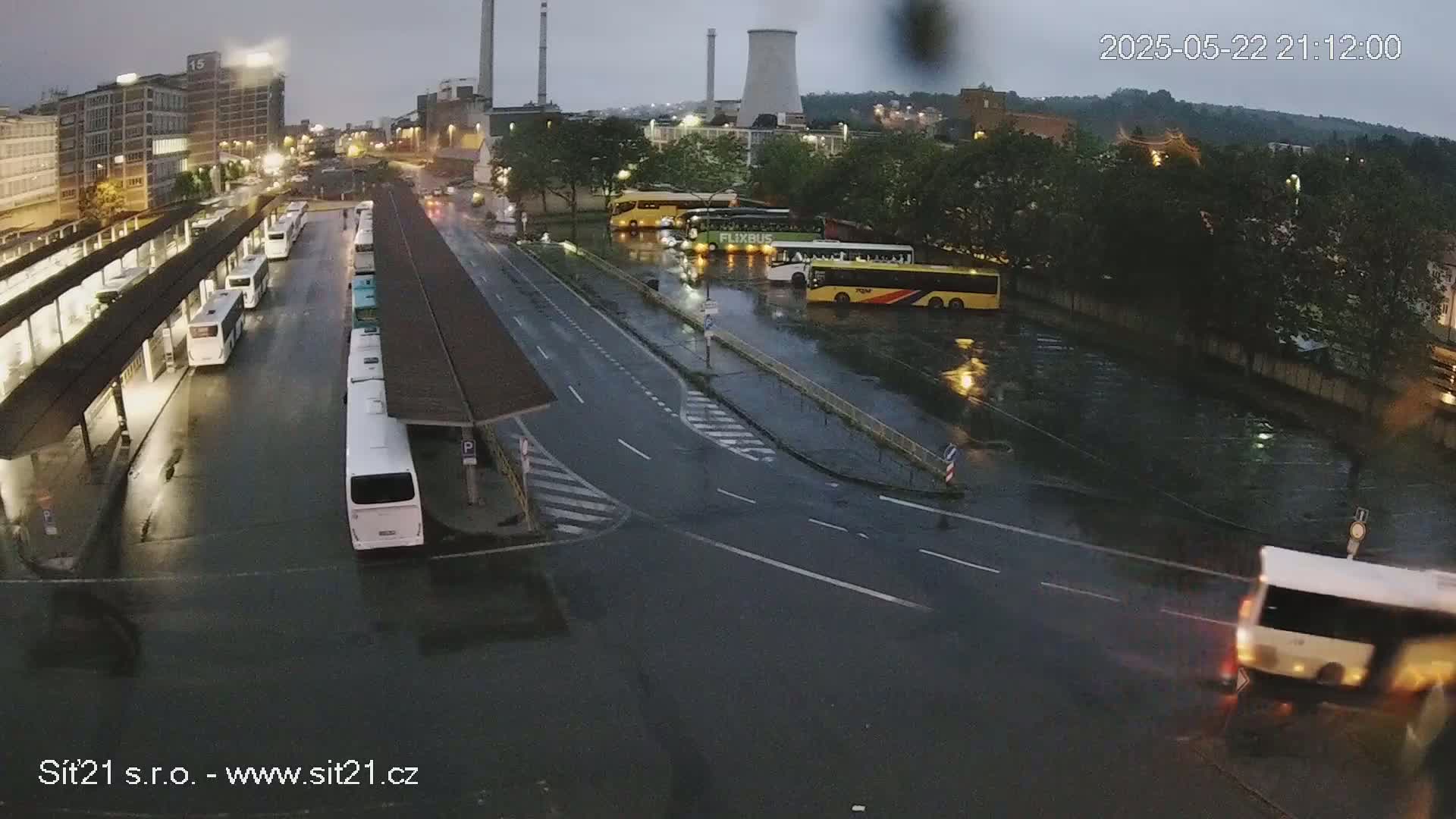 A wet bus station with numerous buses parked and some driving away under an overcast sky.