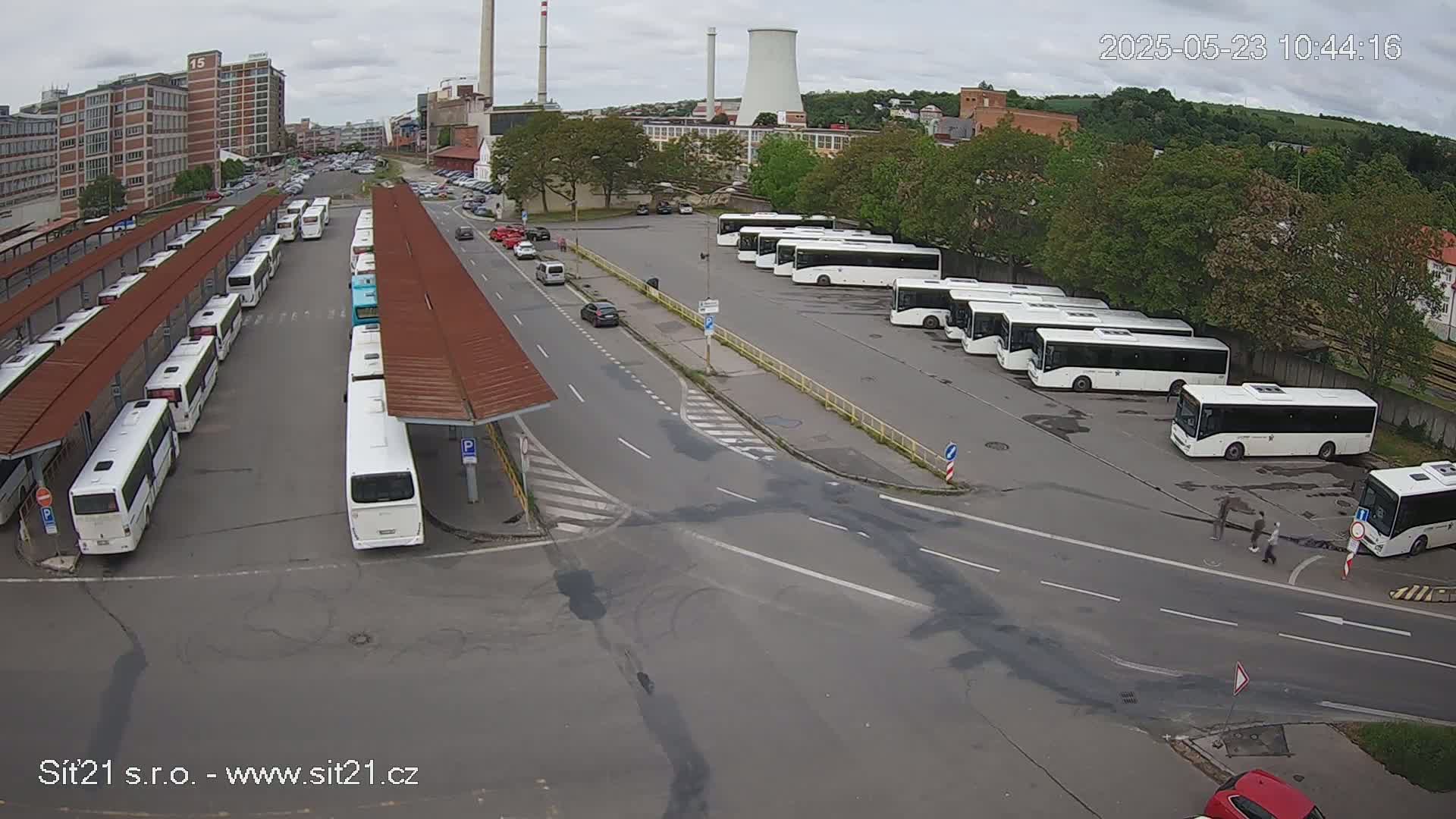 An overcast day reveals a bus station with numerous buses parked under shelters and in a lot, surrounded by roads and industrial buildings.