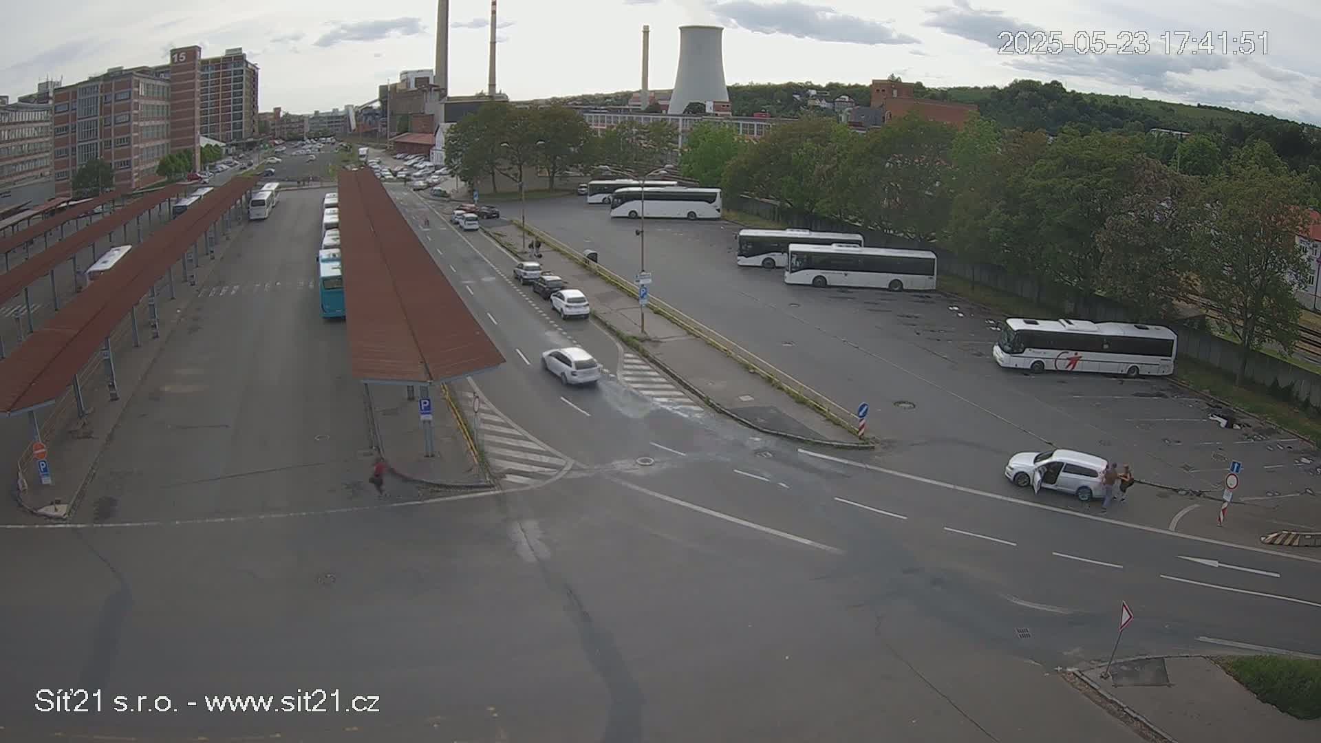 An overcast day shows a bus station with numerous buses parked and a few cars driving on roads around it, near an industrial area with a large cooling tower.