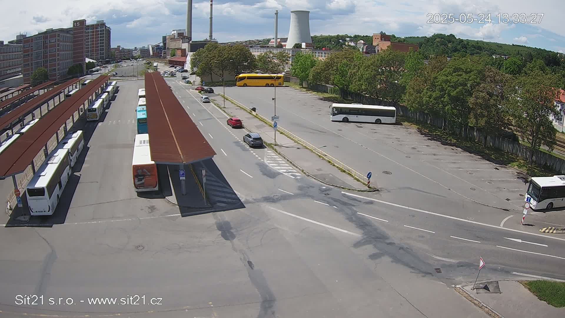 A bus station with numerous buses parked under shelters, several cars in a parking lot, and industrial buildings and trees in the background under a partly cloudy sky.