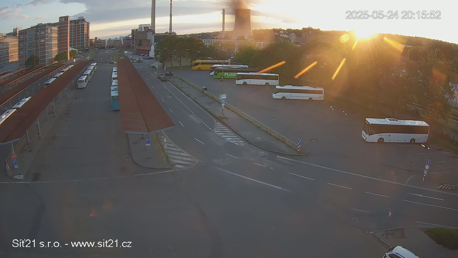 An elevated view of a bus station with several buses parked under covered shelters and in a nearby lot, during a sunny sunset.