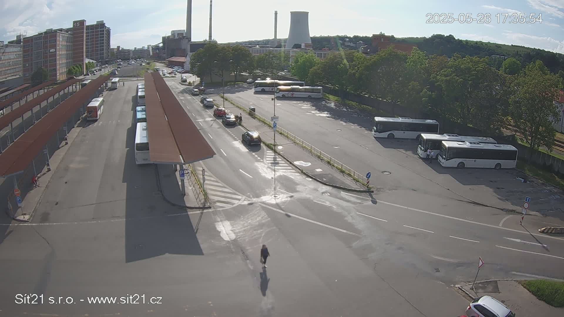 An overhead view of a bus station with several buses parked, cars driving on the roads, and a person walking on a street, under sunny skies.
