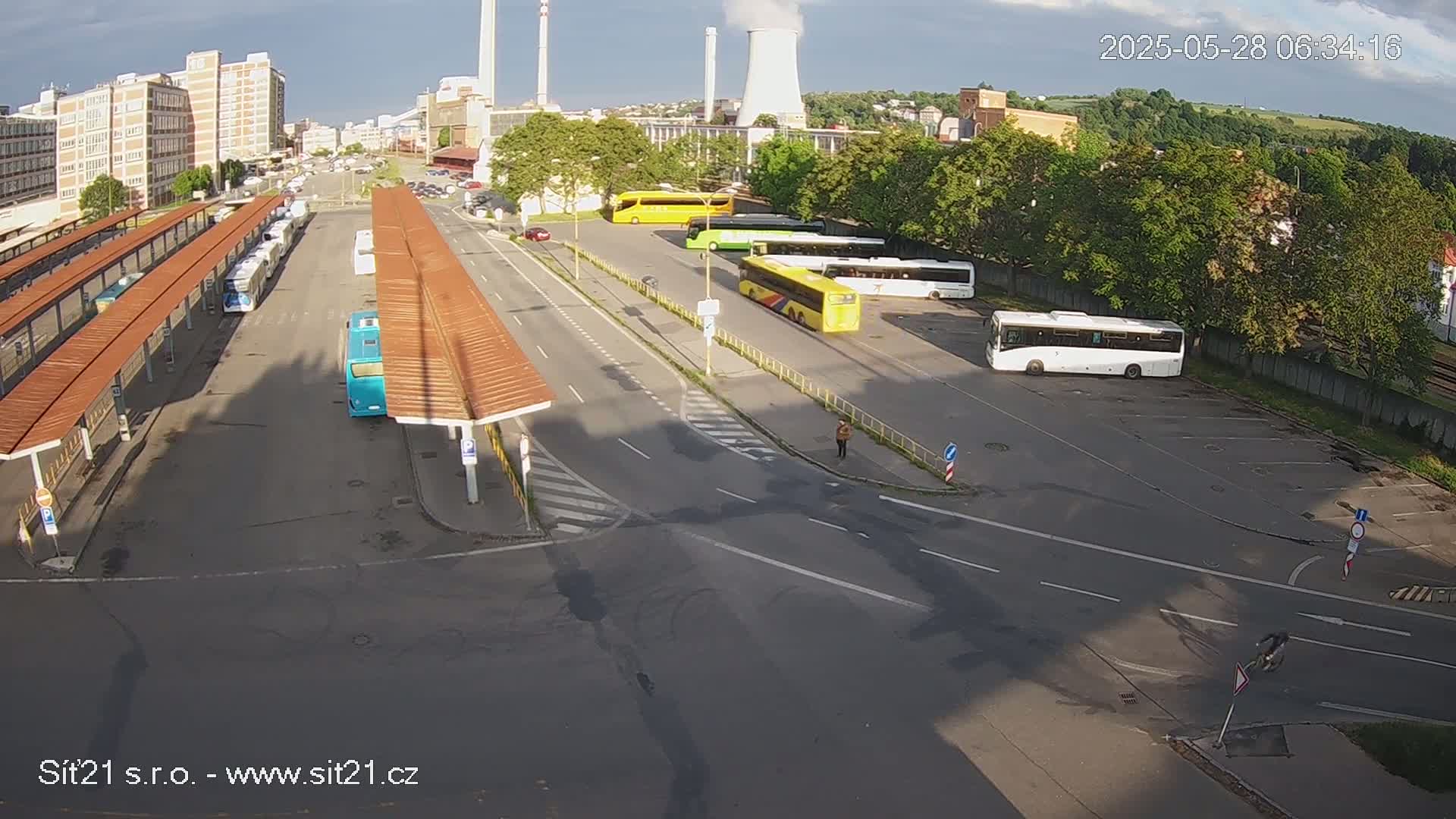 A high-angle view of a bus station with numerous buses parked under partly sunny skies, near industrial buildings and a cooling tower.