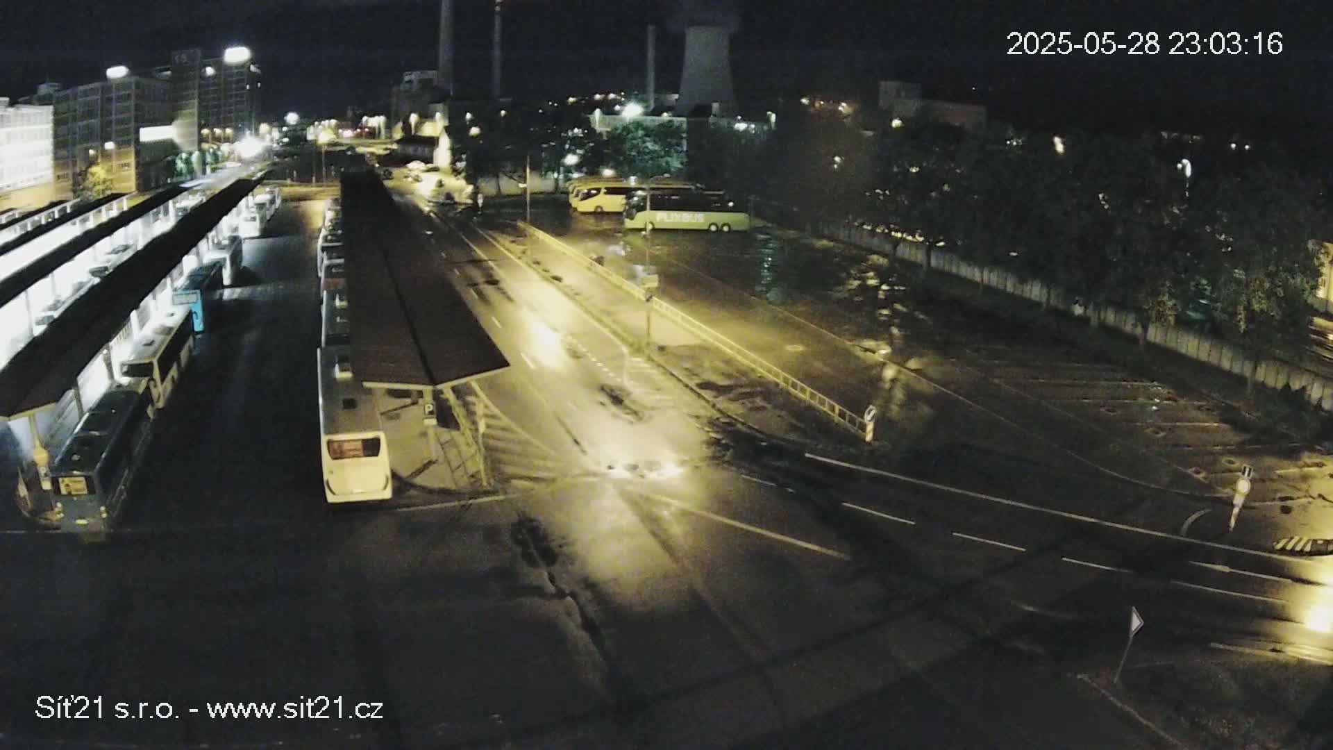 A nighttime, high-angle view of a bus station with several buses parked under covered shelters and in a nearby lot, illuminated by streetlights on wet roads.