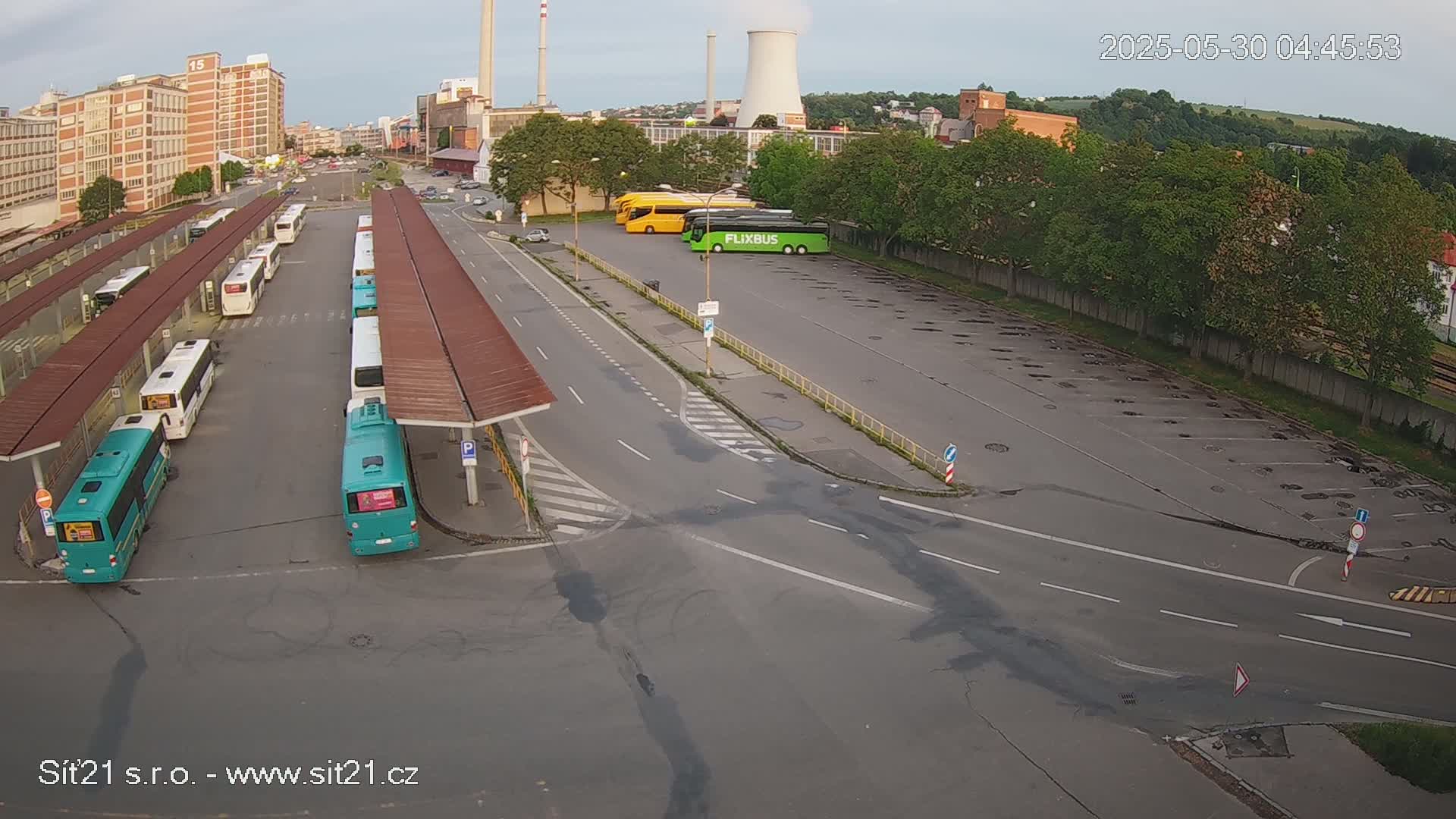 A high-angle view of a bus station with several buses parked under shelters, additional buses parked in a lot nearby, and a mostly clear sky.