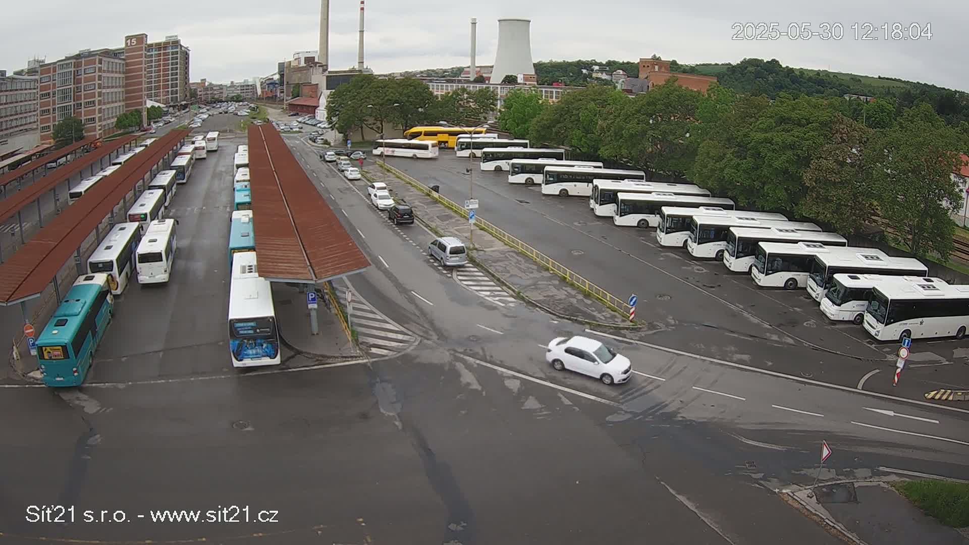 A bus station with numerous buses parked under shelters, along with several cars in a parking lot under an overcast sky.