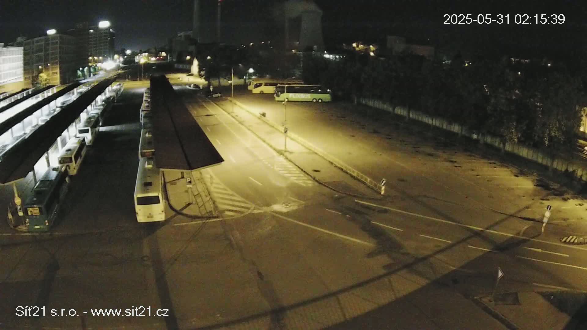 A nighttime, high-angle view shows a bus station with numerous buses parked under shelters, and several more buses parked in a lot across a roadway.