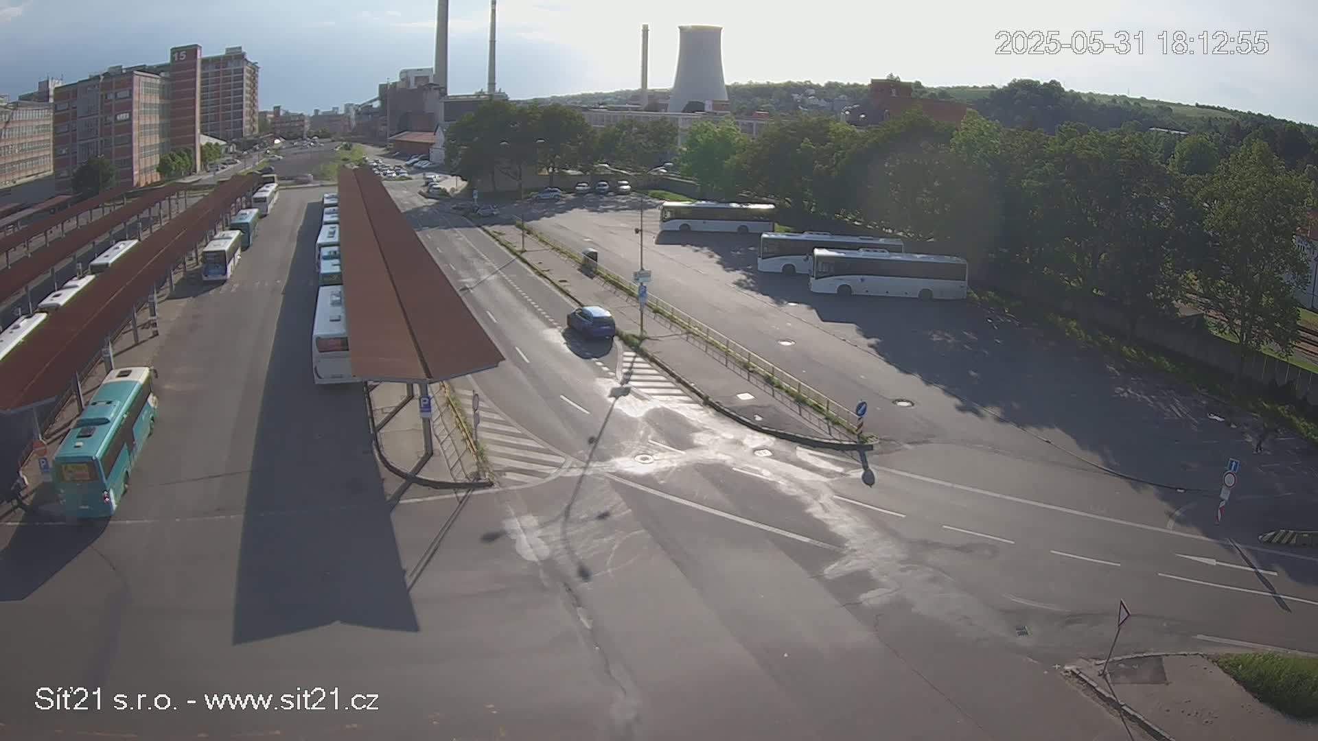 A sunny overhead view shows a bus station with numerous buses parked under shelters, a roadway intersecting the station, and a parking lot with additional buses and a single car, all situated near industrial buildings and greenery.