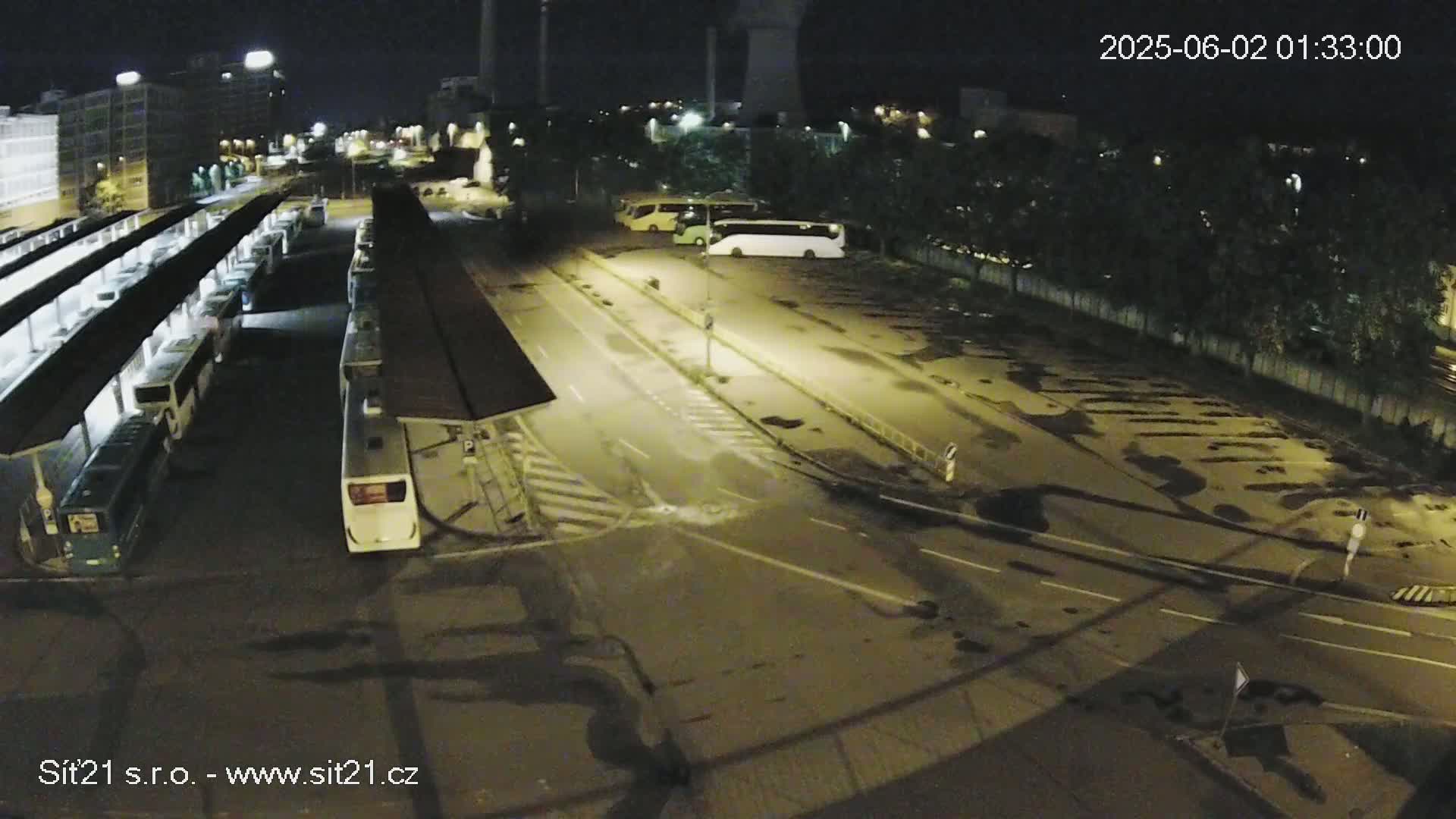 A nighttime view of a bus station with numerous buses parked under covered shelters and additional buses parked in a nearby lot, under partly cloudy skies.