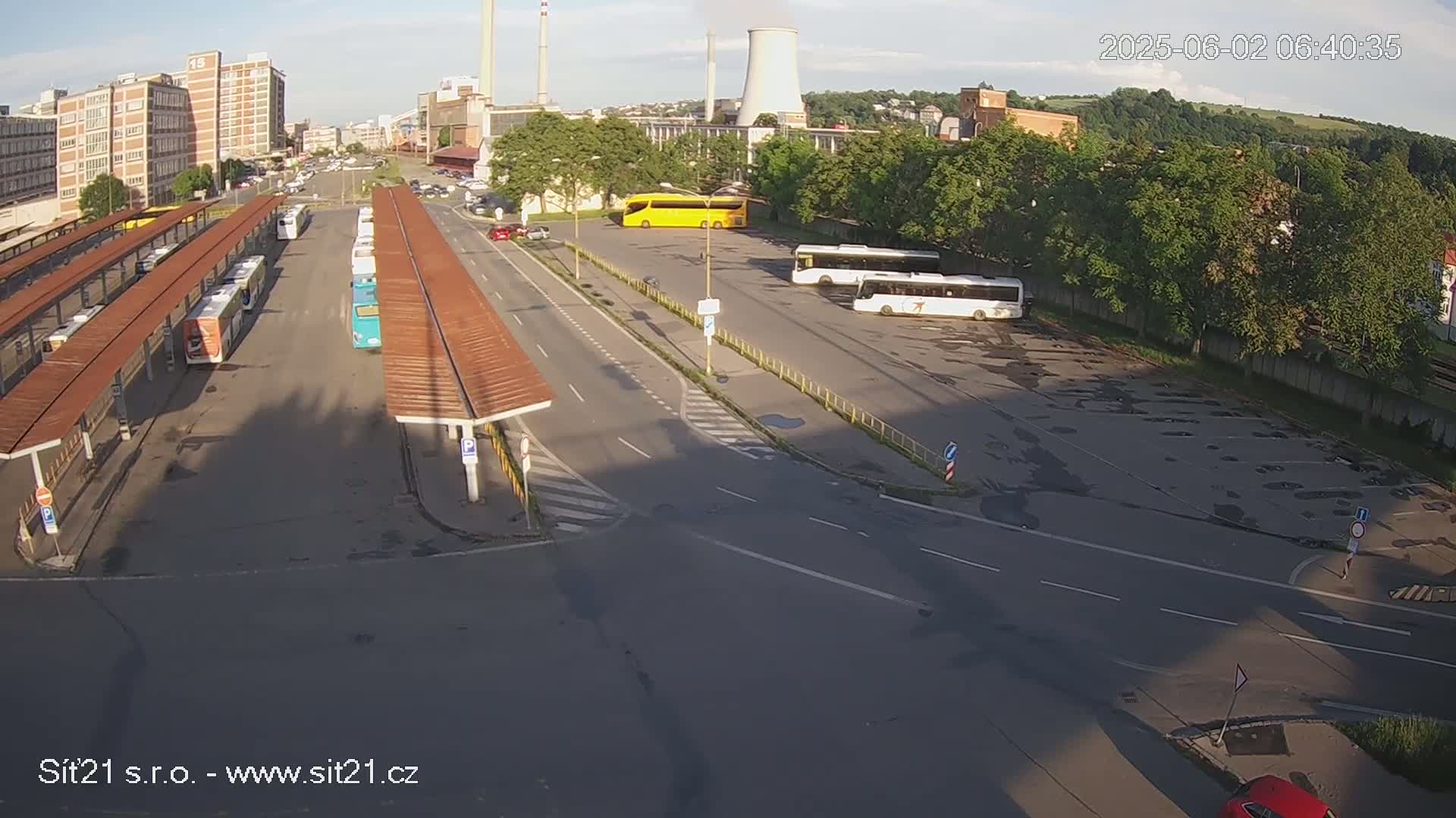 An aerial view shows a bus station with numerous buses parked under shelters and in a lot, alongside a roadway, under a partly sunny sky.