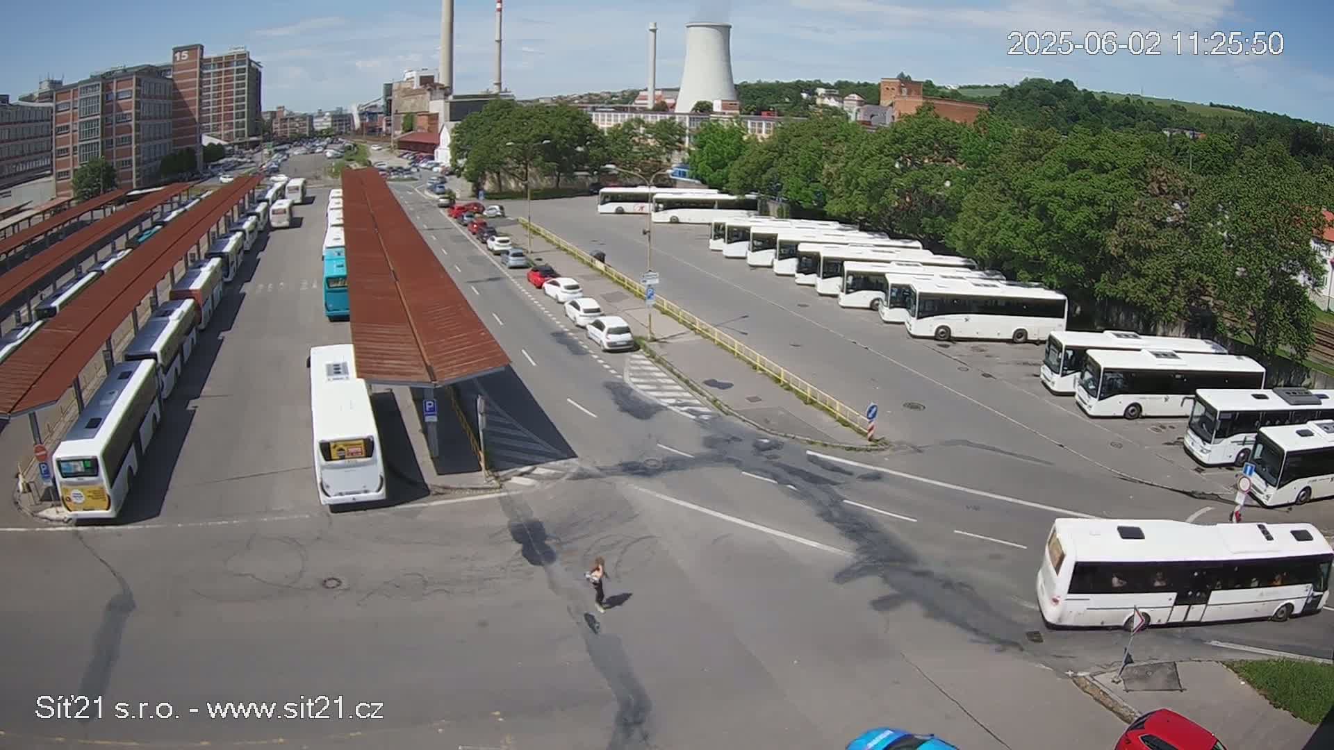 A sunny day reveals a bus depot with numerous buses parked, some under covered shelters, and a few cars on surrounding roads.