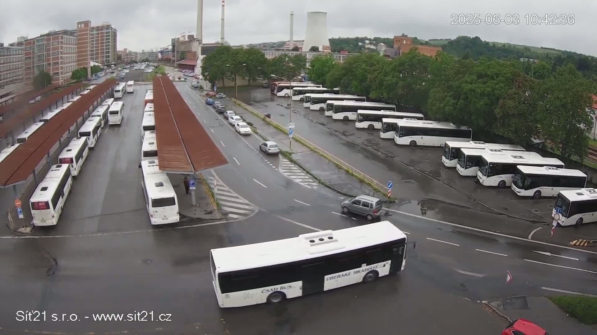 A bus depot with numerous white buses parked under covered areas, with several cars on the surrounding wet roads under an overcast sky.