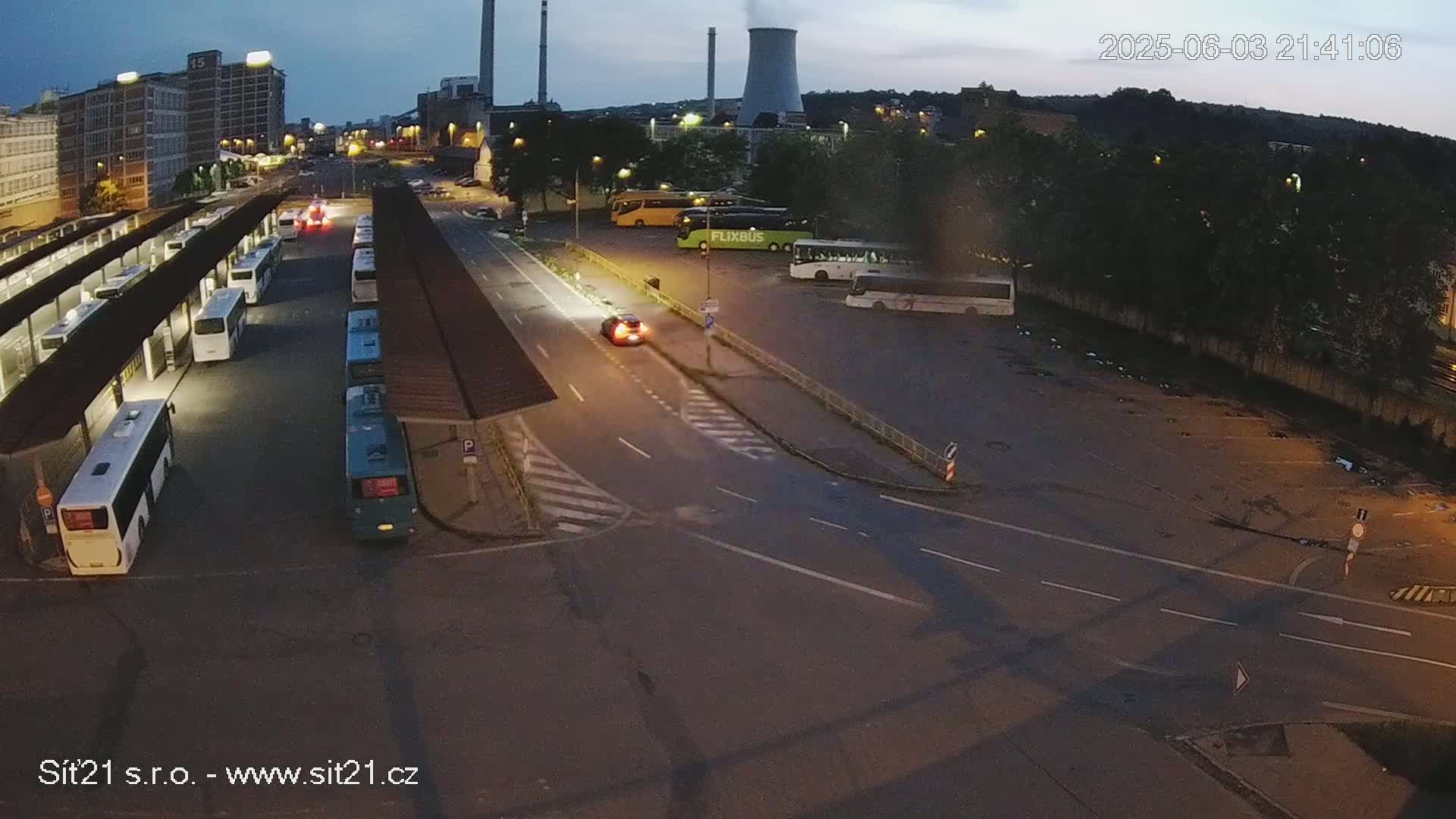 A high-angle, nighttime view shows a bus station with several buses parked under shelters, a car driving on a road, and a large empty parking lot next to a factory under a twilight sky.