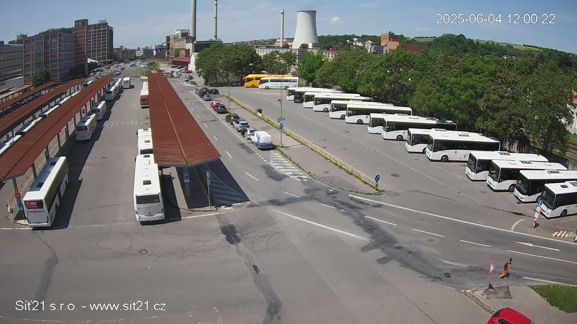 A bus station with numerous buses parked under covered shelters on a sunny day, with additional buses parked in an open lot nearby.