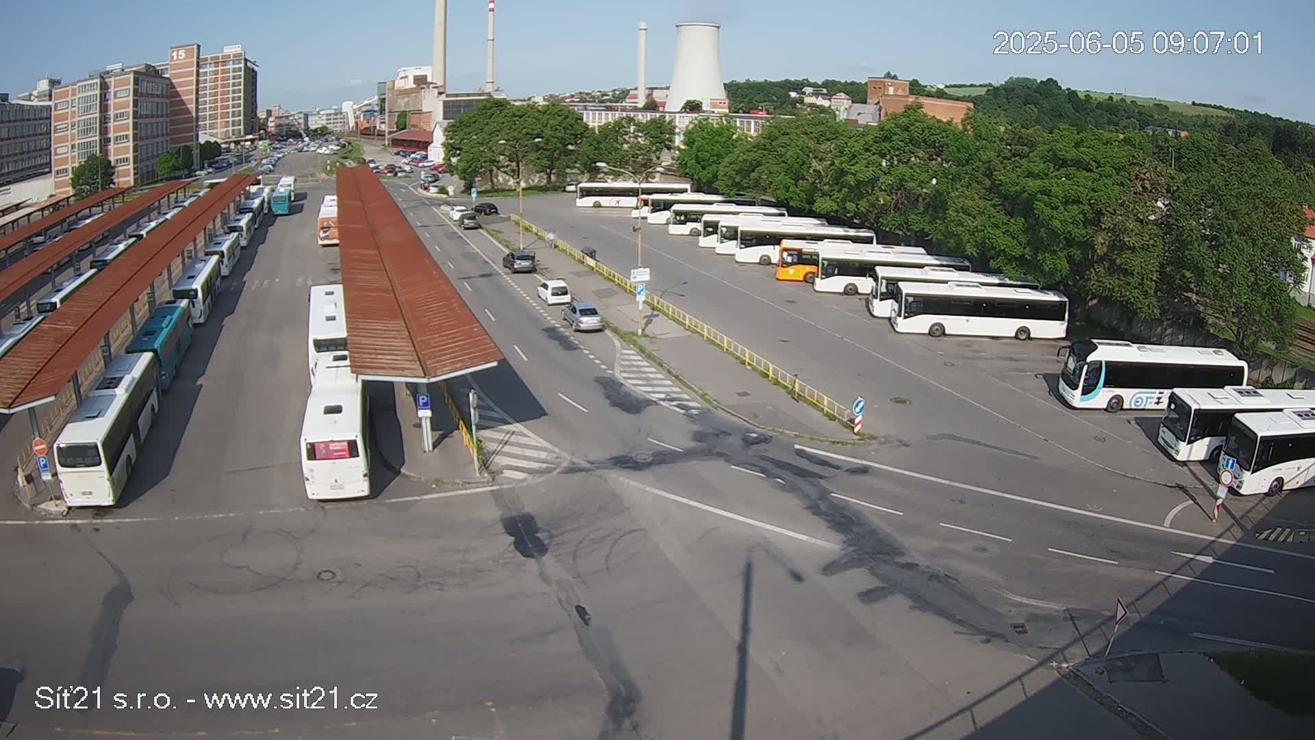 A sunny day shows a bus depot with many buses parked under shelters and in a lot, along with several cars on adjacent roads and industrial buildings in the background.