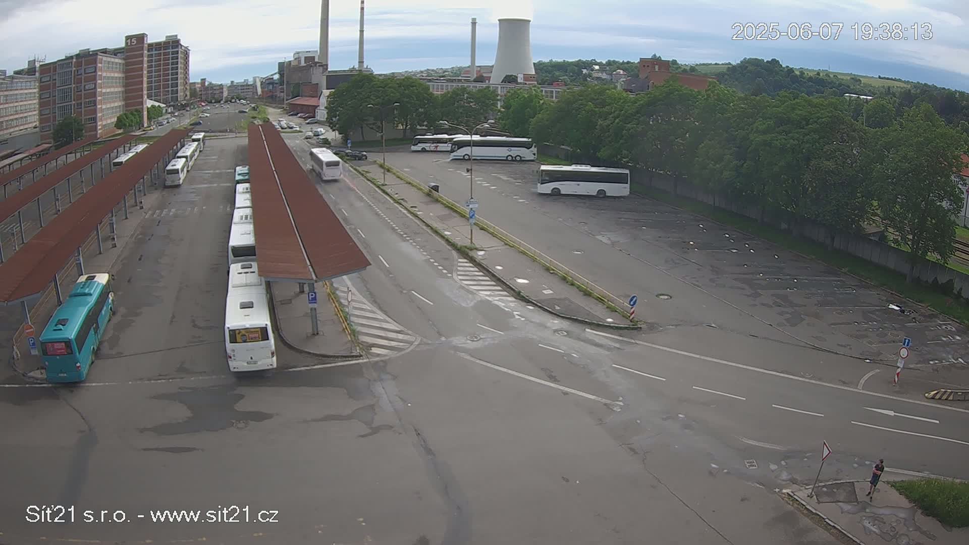 A bus station with several buses parked under shelters and in a nearby lot, situated near industrial buildings under an overcast sky.
