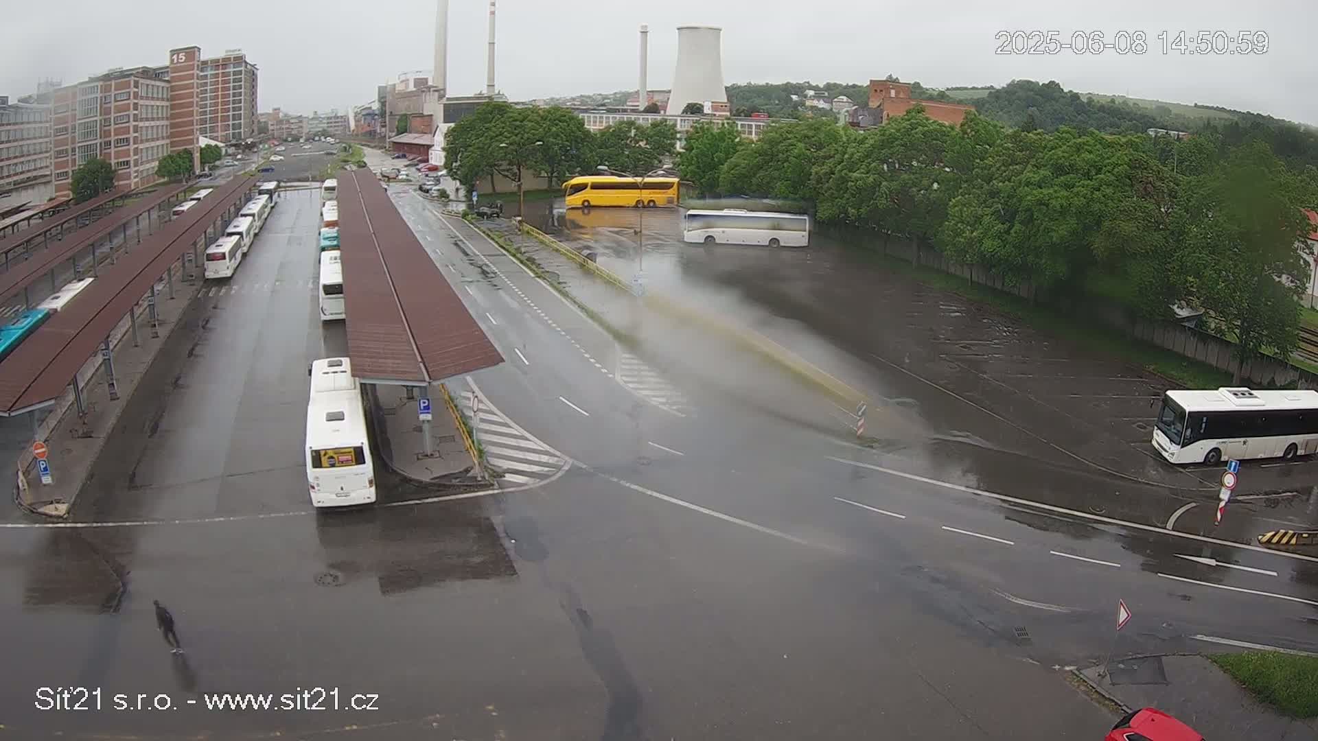 A wet bus station with numerous buses parked under covered shelters, surrounded by roadways and industrial buildings under an overcast sky.