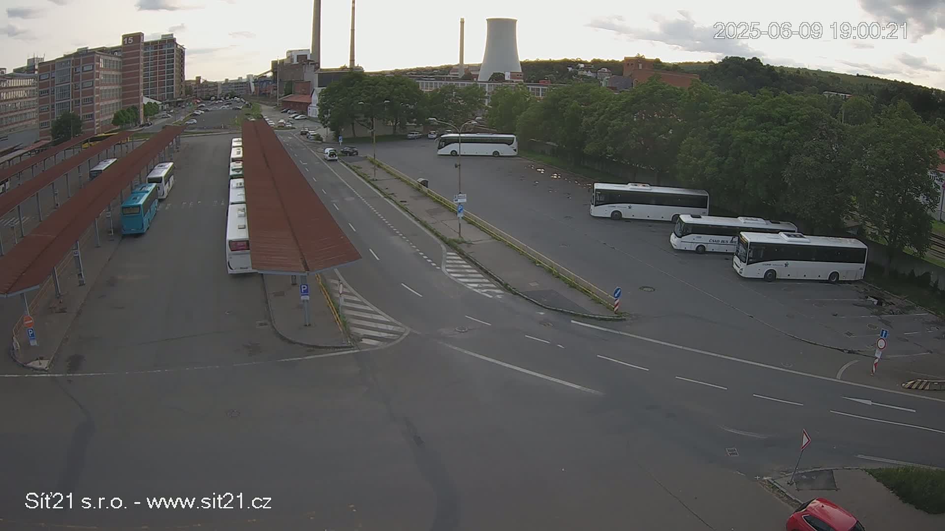An overcast day shows a bus station with numerous buses parked at covered and uncovered areas, near an industrial area with a large cooling tower.