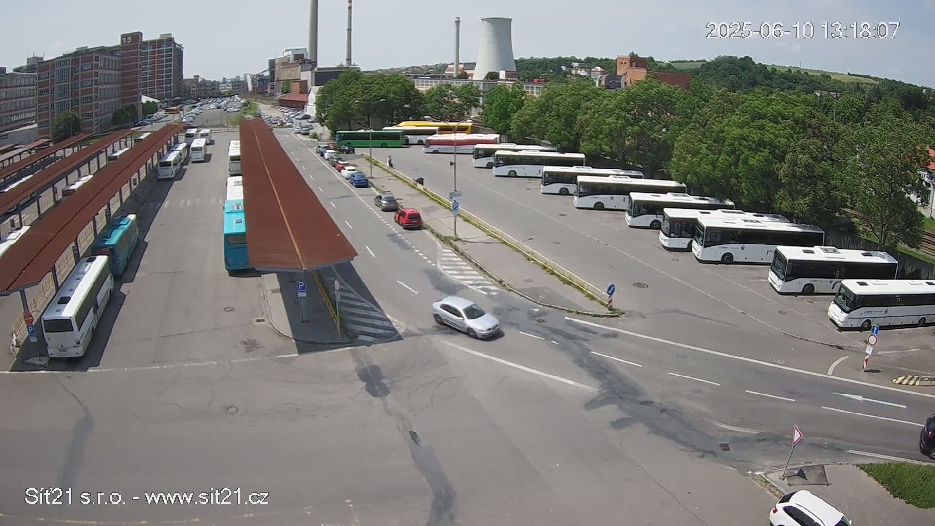 A bus station with numerous buses parked under covered shelters and several cars driving on roads nearby on a sunny day.