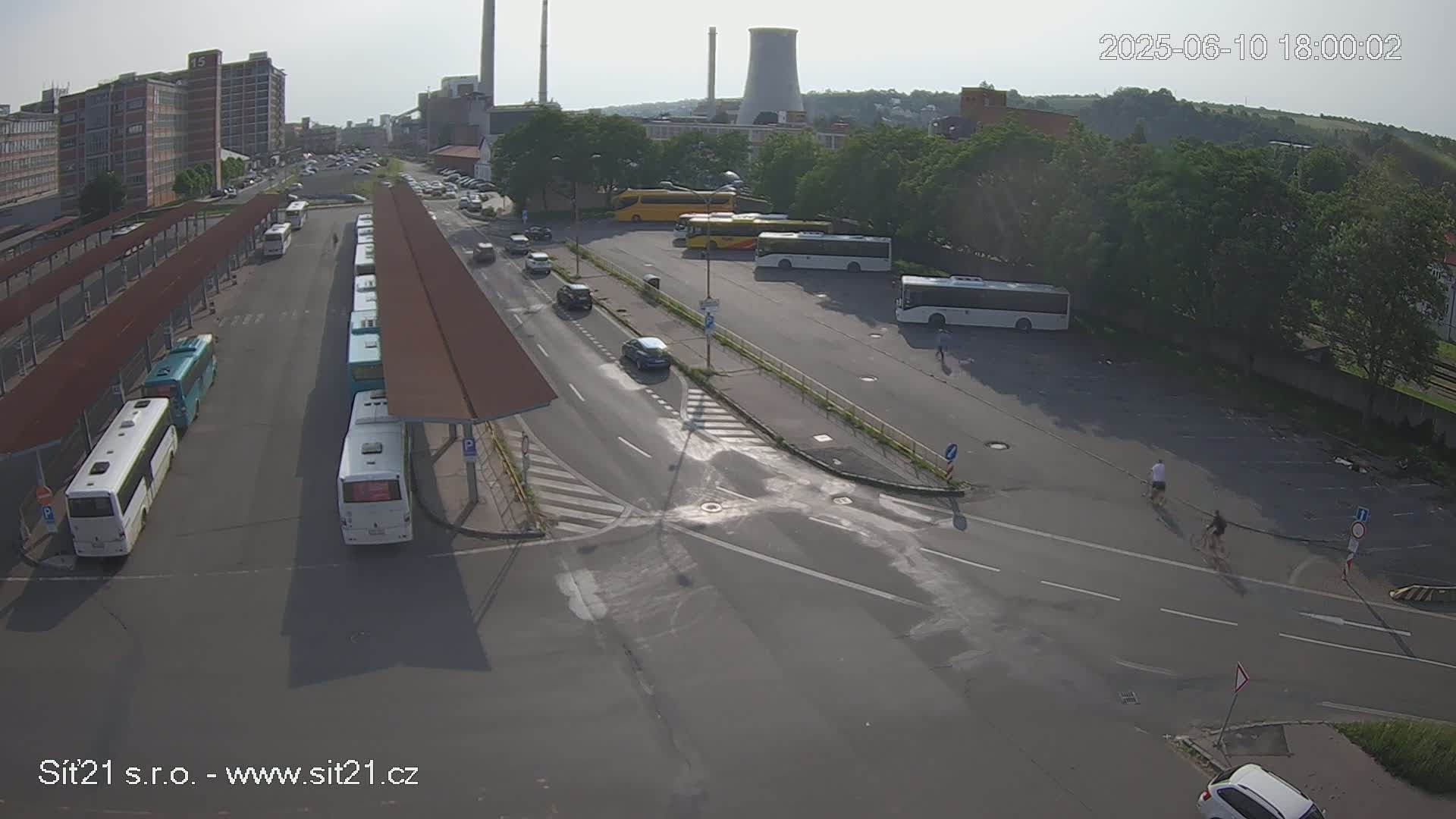A high-angle, sunny view of a bus station with numerous buses parked under shelters, several cars driving on adjacent roads, and a few people walking and biking nearby, with industrial buildings in the background.
