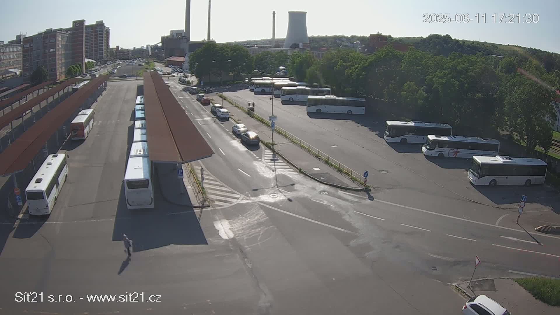 A high-angle, sunny view of a bus station with numerous buses parked, some cars driving by, and a cooling tower in the background.