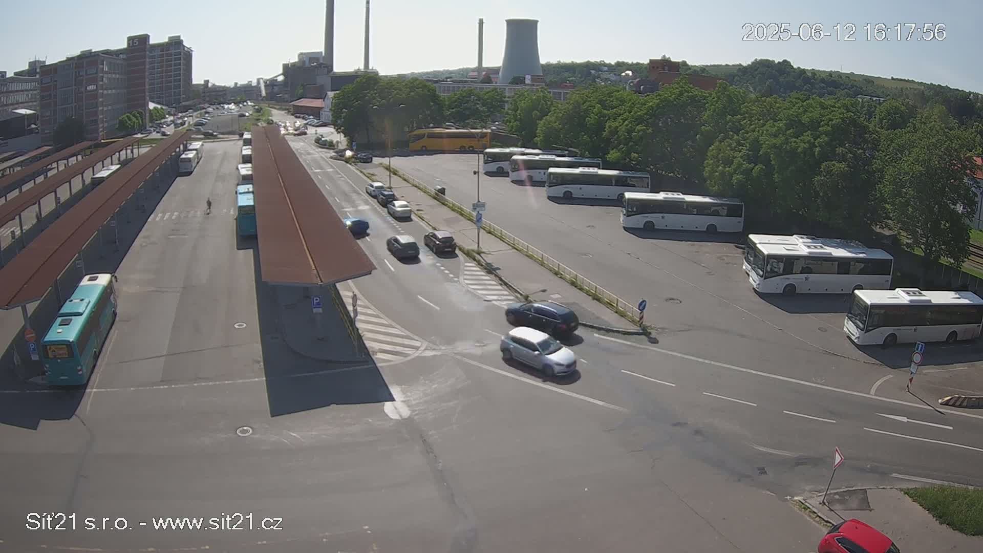 A high-angle, sunny view of a bus station with numerous buses parked, several cars driving on the surrounding roads, and buildings in the background.