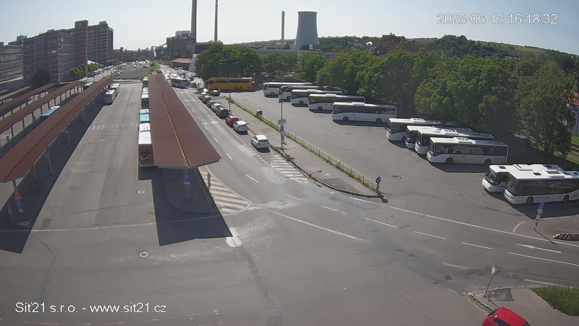 A bus station with numerous buses parked under a sunny sky, next to industrial buildings and a cooling tower in the background.