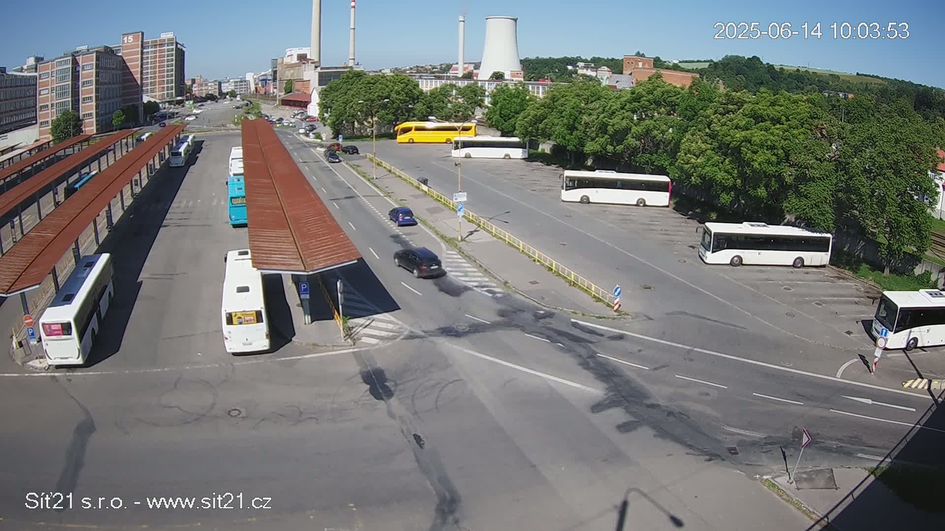 A sunny day at a bus station with numerous buses parked under covered areas and in an open lot, alongside a road with a few cars.