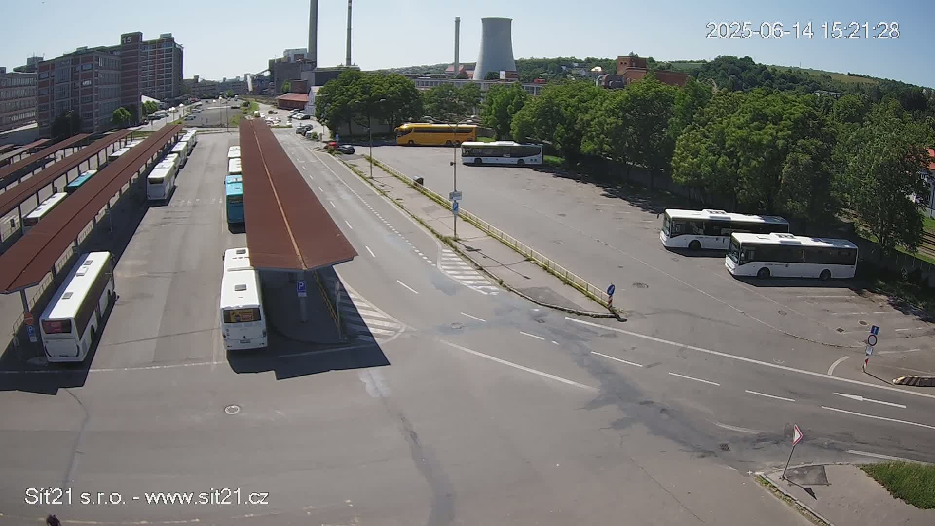 A high-angle, sunny-day view of a bus station with numerous buses parked under shelters and in a nearby lot, next to industrial buildings and green trees.