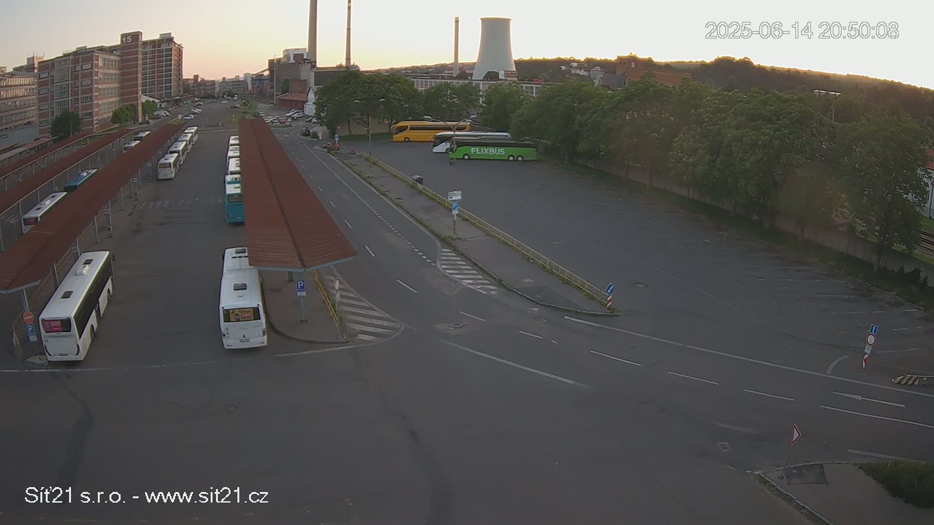A high-angle, sunny-day view of a bus station with numerous buses parked under shelters and in a nearby lot, next to industrial buildings and green trees.