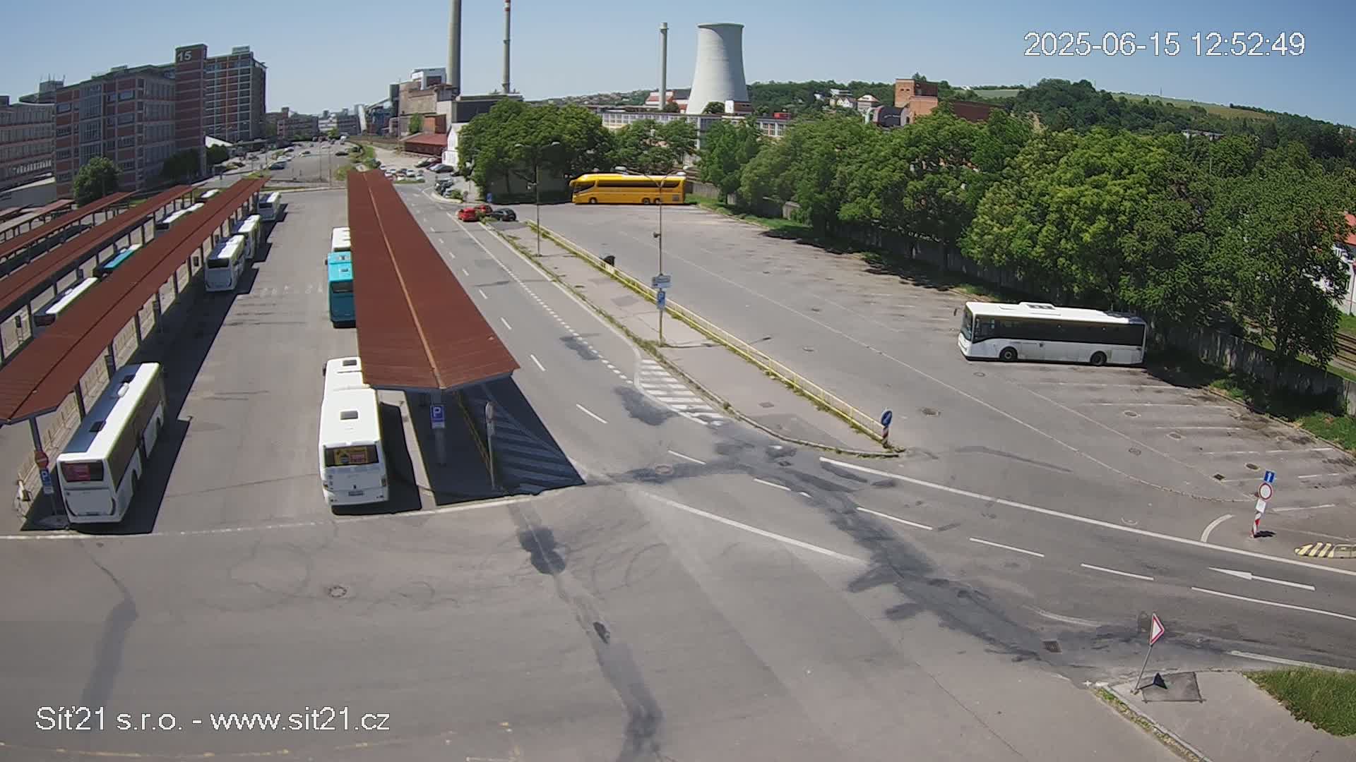 A bus station with several buses parked under shelters, a large empty paved lot, and a road intersection on a sunny day.