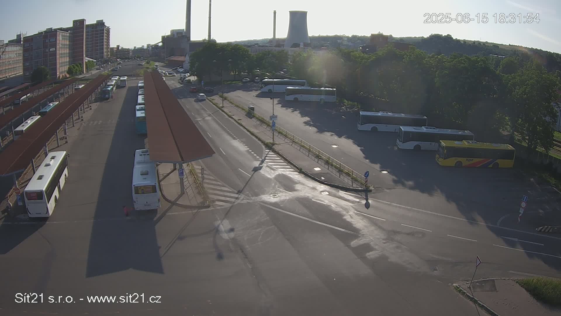 A sunny day shows a bus station with numerous buses parked under covered shelters and in a nearby lot, adjacent to a road intersection near industrial buildings and a cooling tower.