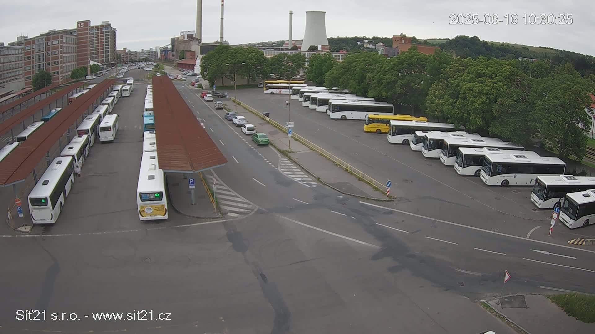 An overcast day shows a bus station with many buses parked at the platforms and in a lot, with a few cars on the road nearby.