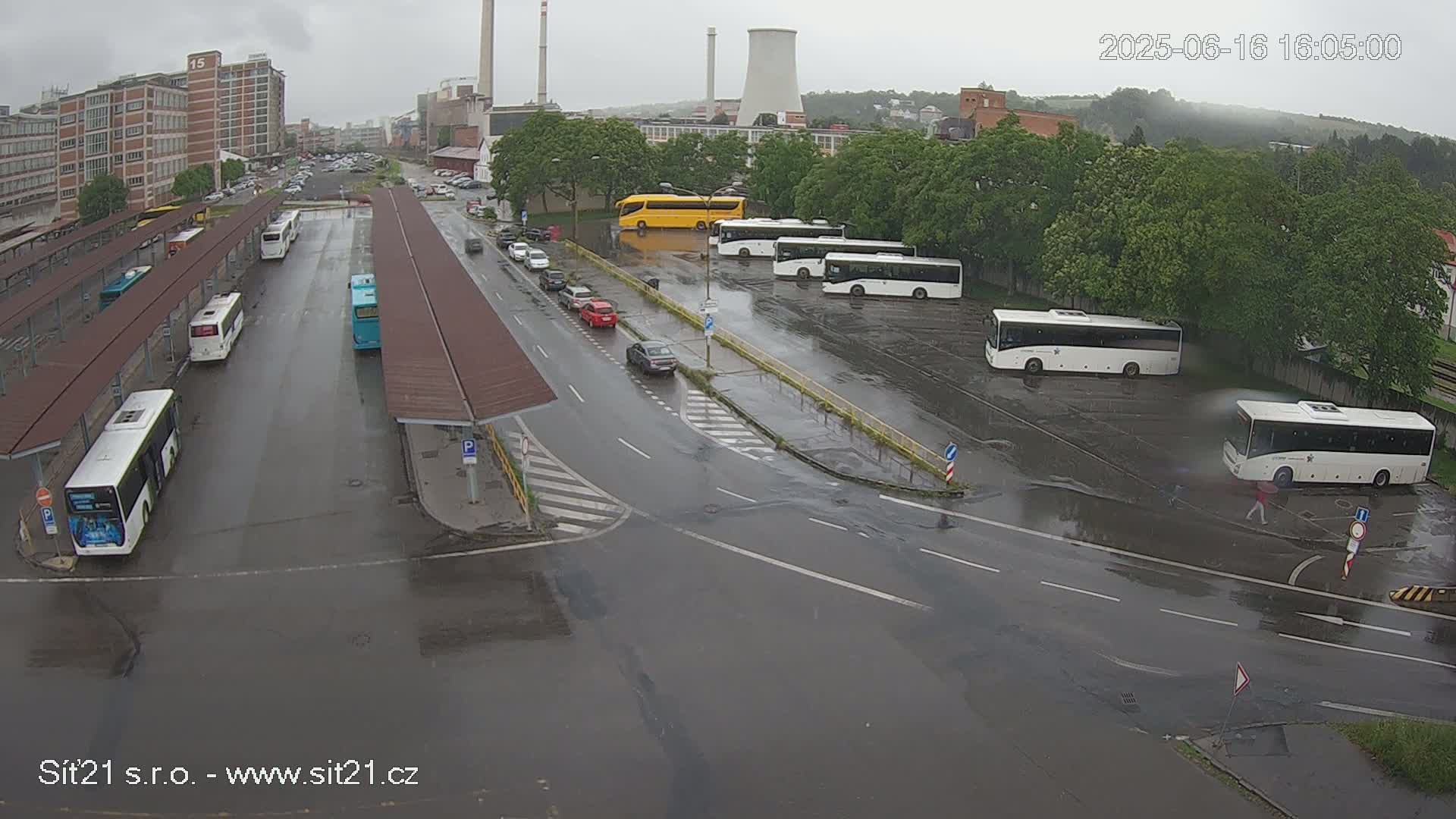 A wet bus station with numerous buses parked under covered shelters and in an adjacent lot, during a rainy day.
