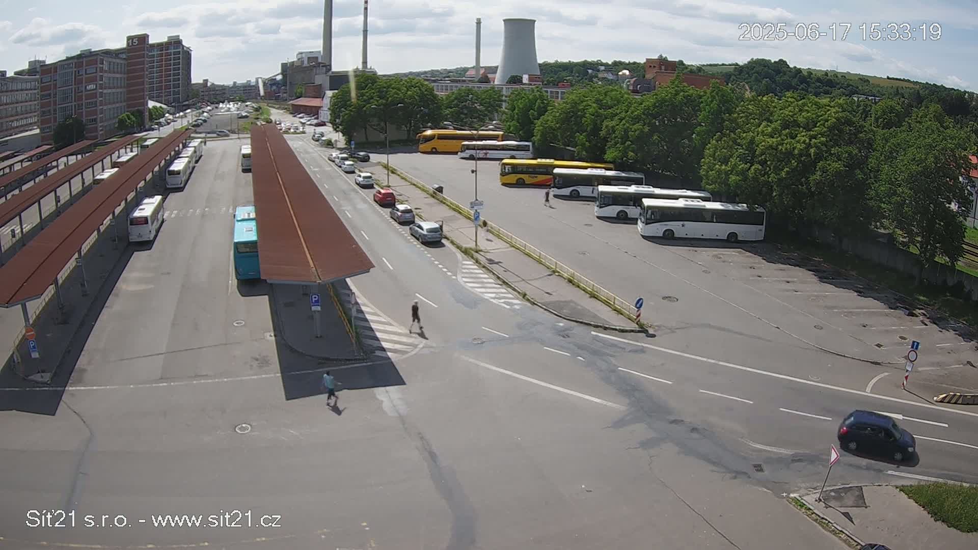 A bus station with numerous buses parked under covered shelters, several cars on nearby roads, and a cooling tower in the background under a partly sunny sky.