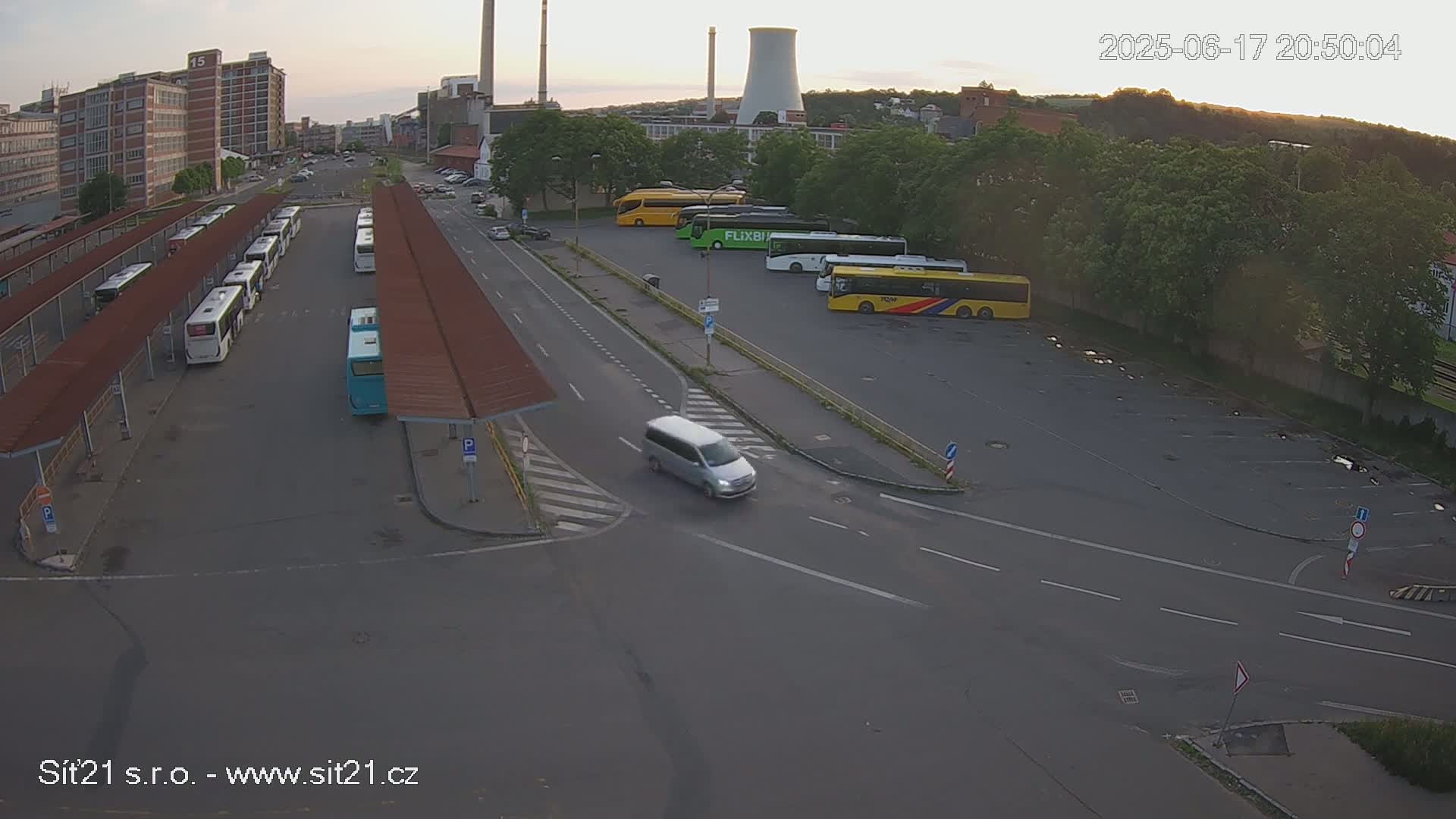 A bus station with numerous buses parked under shelters, along with additional buses and a van in a nearby lot, during the evening under a partly cloudy sky.