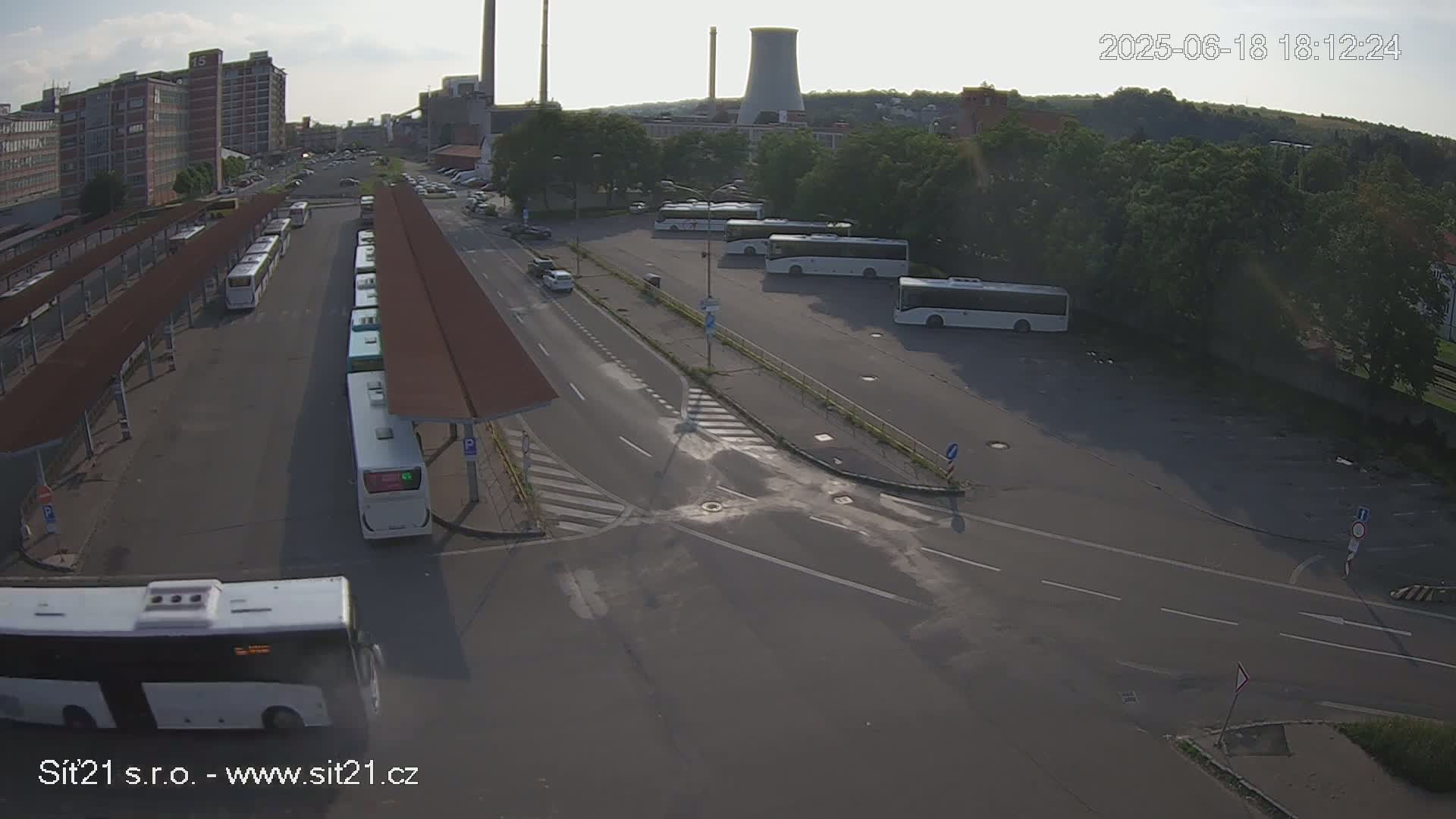 An aerial view of a bus station with numerous buses parked under covered shelters on a sunny day, next to a road intersection.