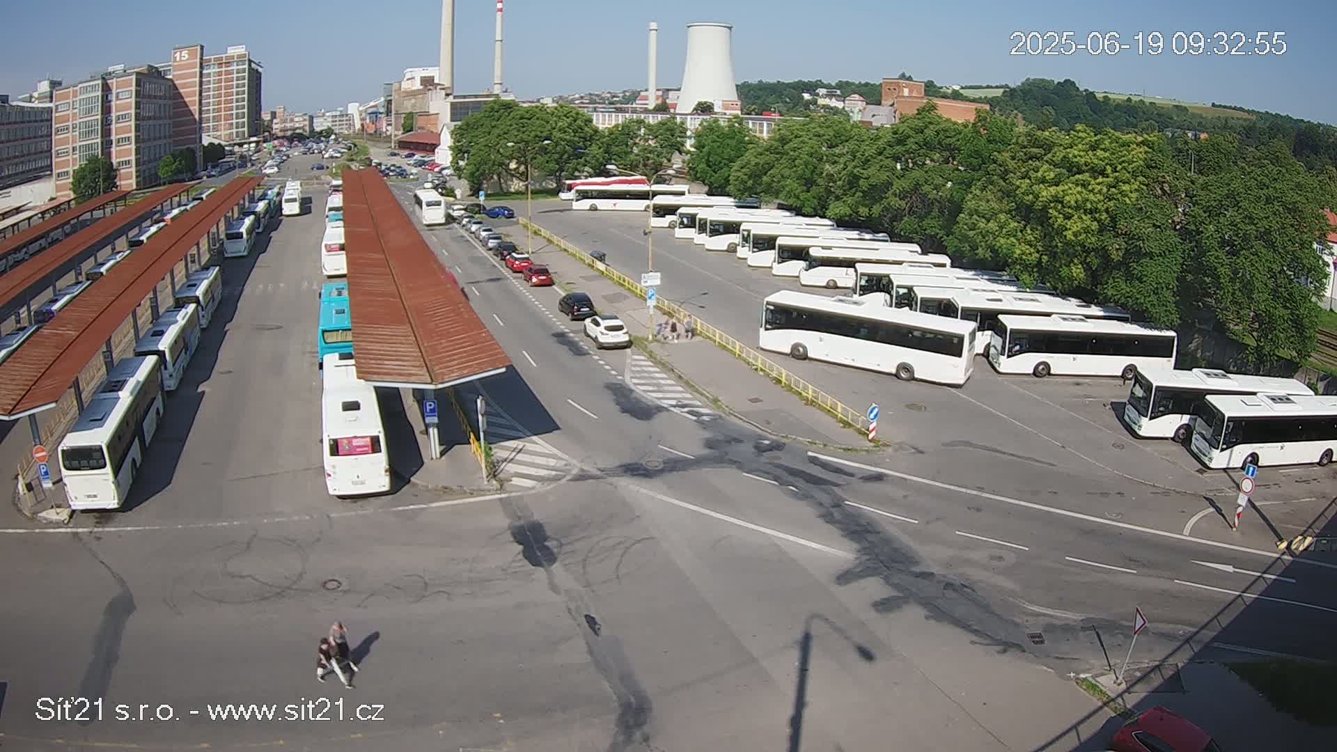 A bus station with numerous buses parked under shelters on a sunny day, alongside a road with cars and a few pedestrians.
