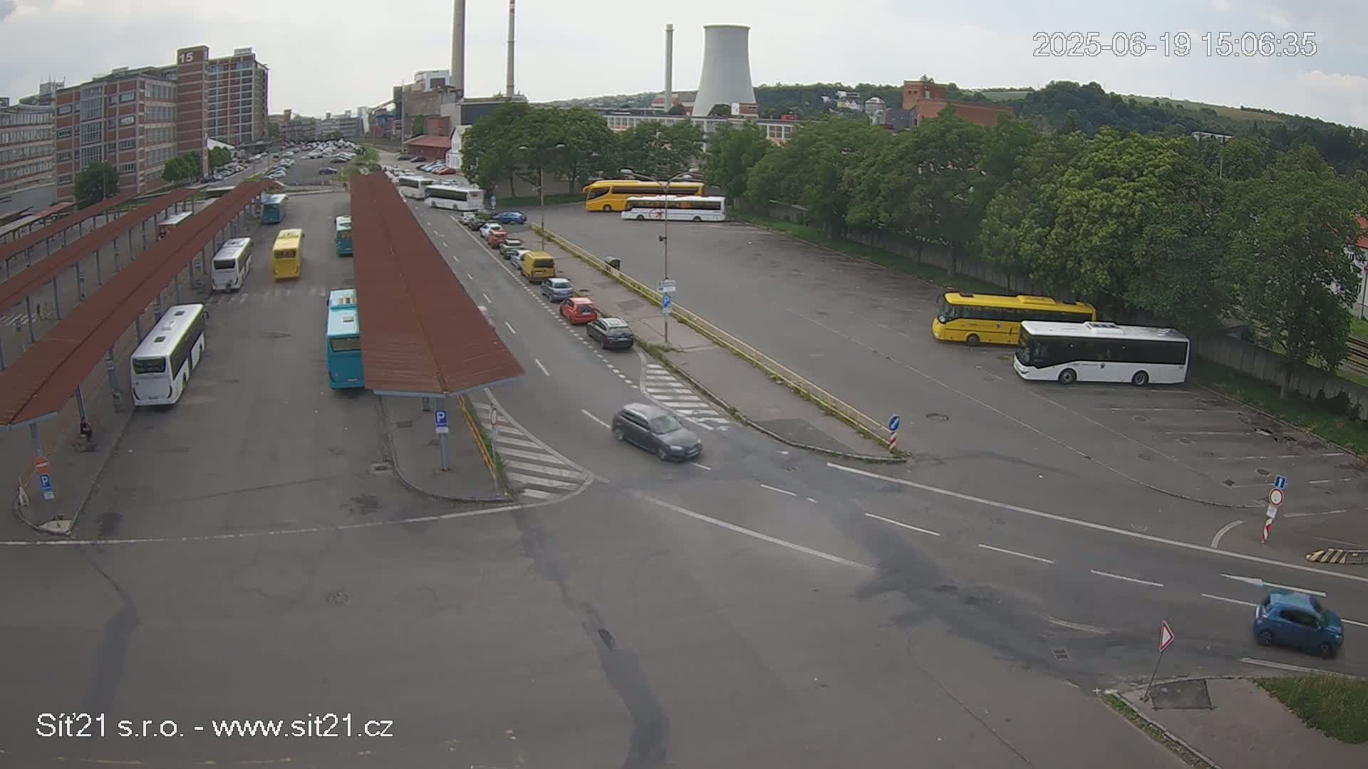 An overcast day shows a bus station with multiple buses parked under shelters and in a nearby lot, along with several cars driving and parked on surrounding roads.
