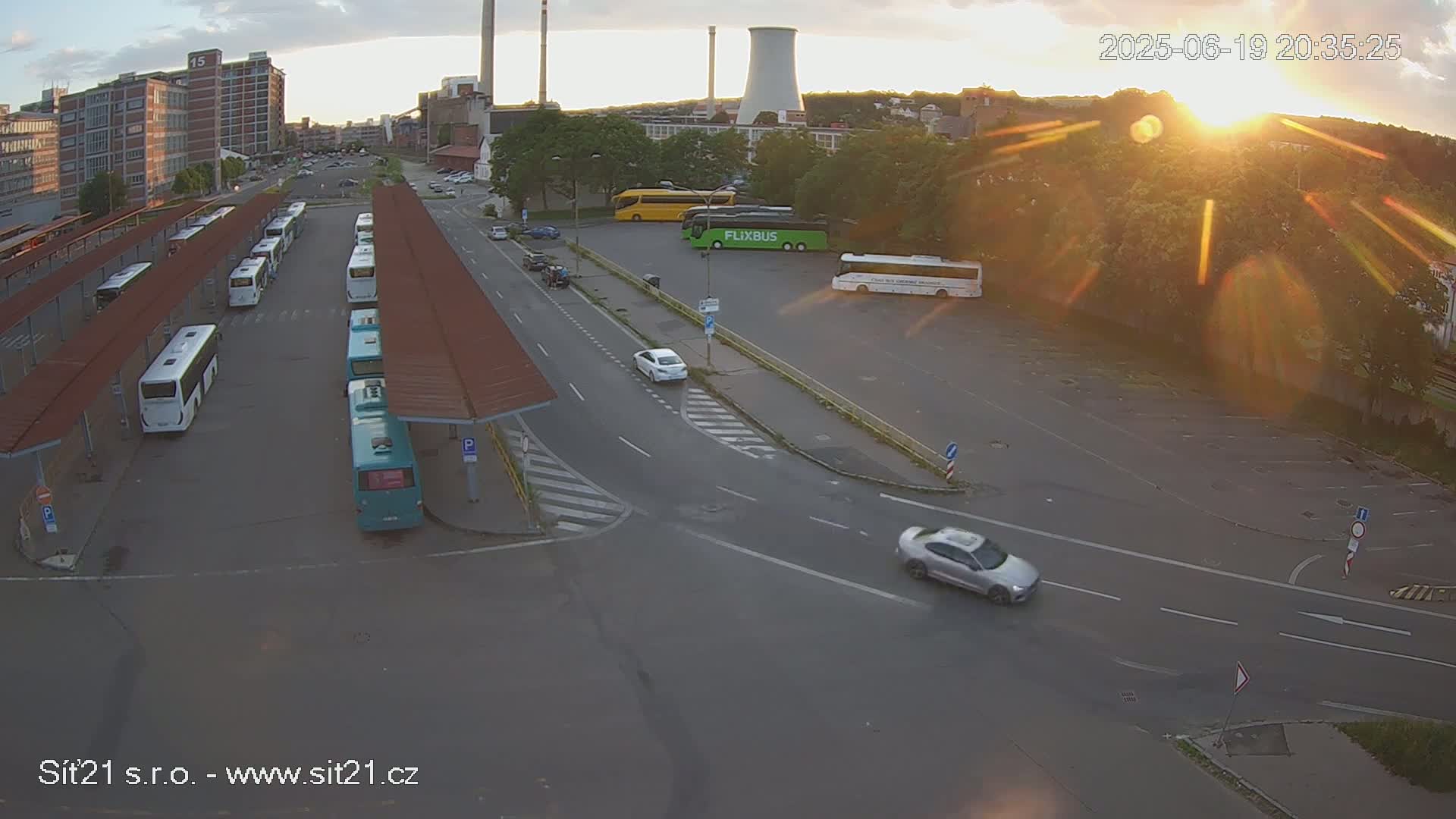 A high-angle view of a bus station and surrounding area at sunset, with numerous buses parked under shelters, cars driving on roads, and a large industrial structure in the background.