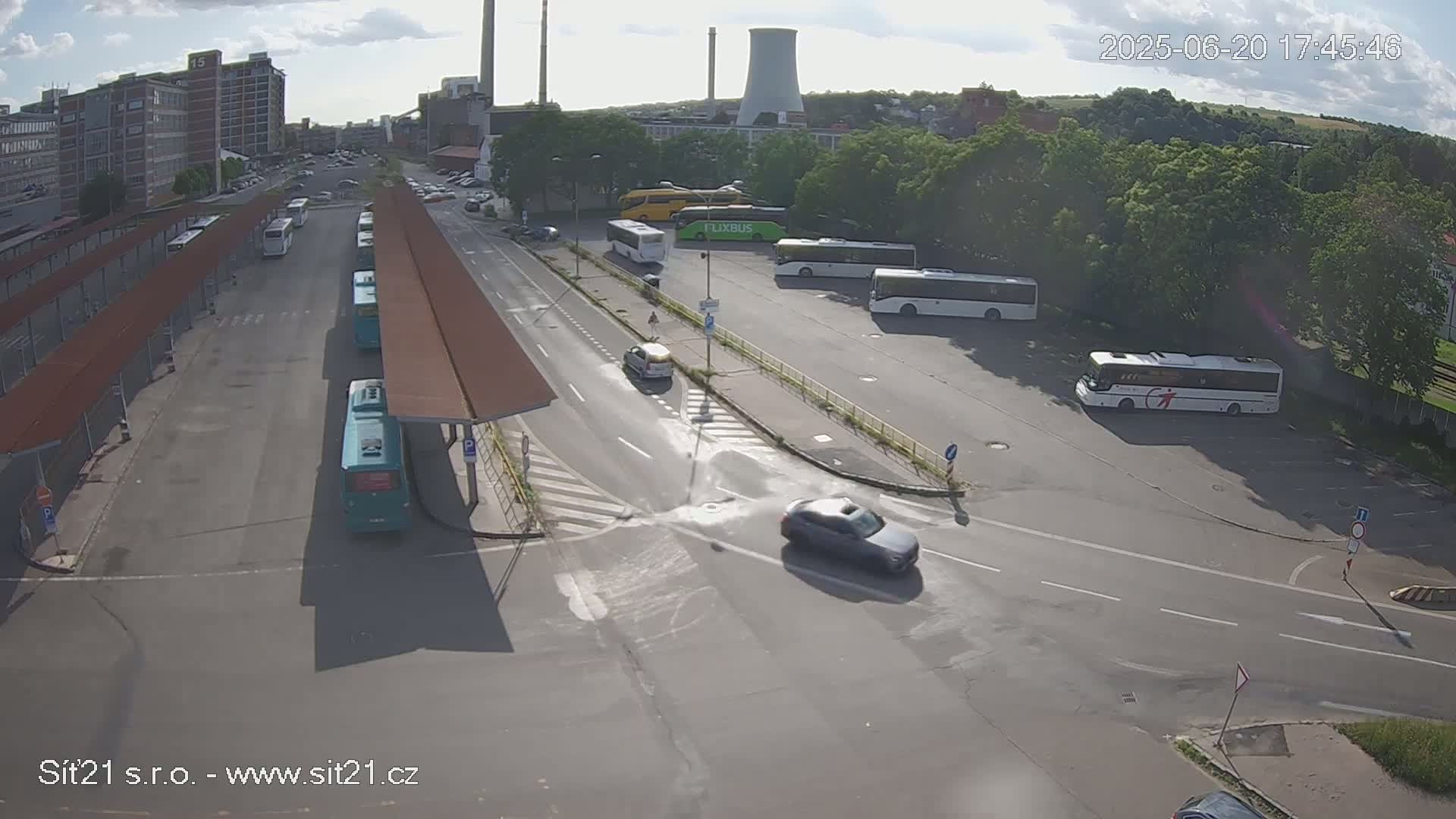 A bus station with numerous buses parked under shelters and in a nearby lot, with several cars driving on surrounding roads, under mostly sunny skies.